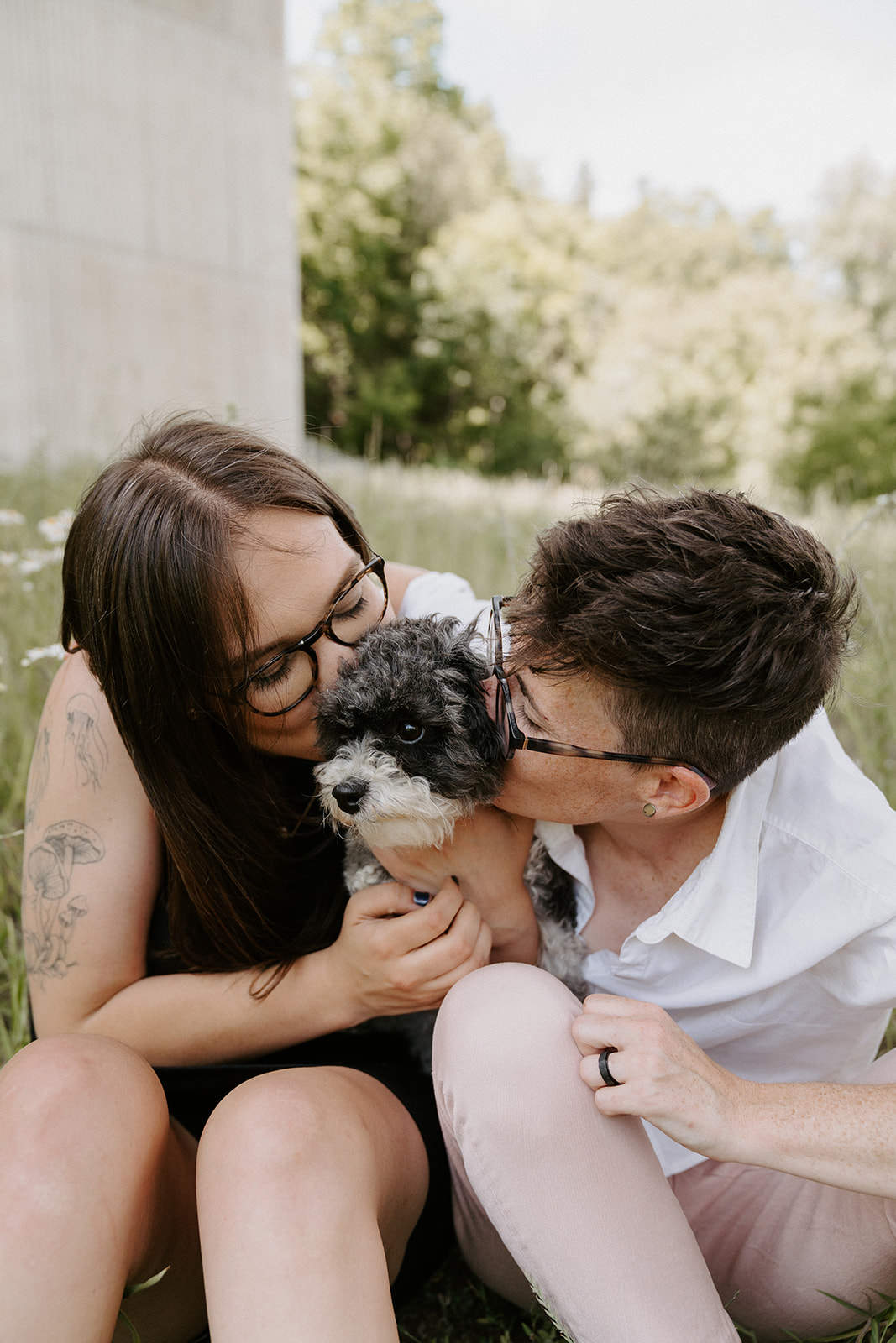 Two people sitting on the grass both kissing the dog between them.