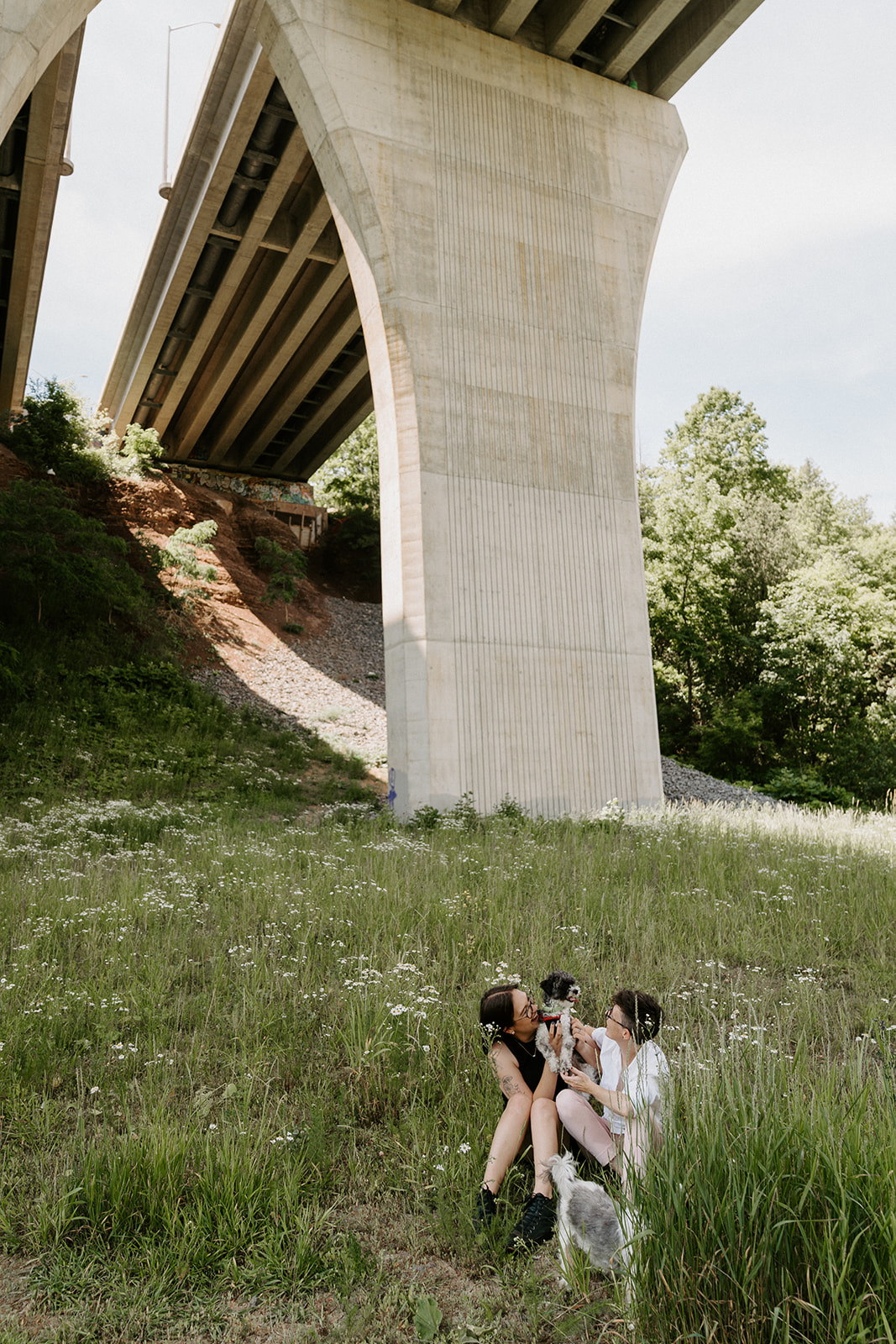 Two people sitting on the grass underneath an overpass.