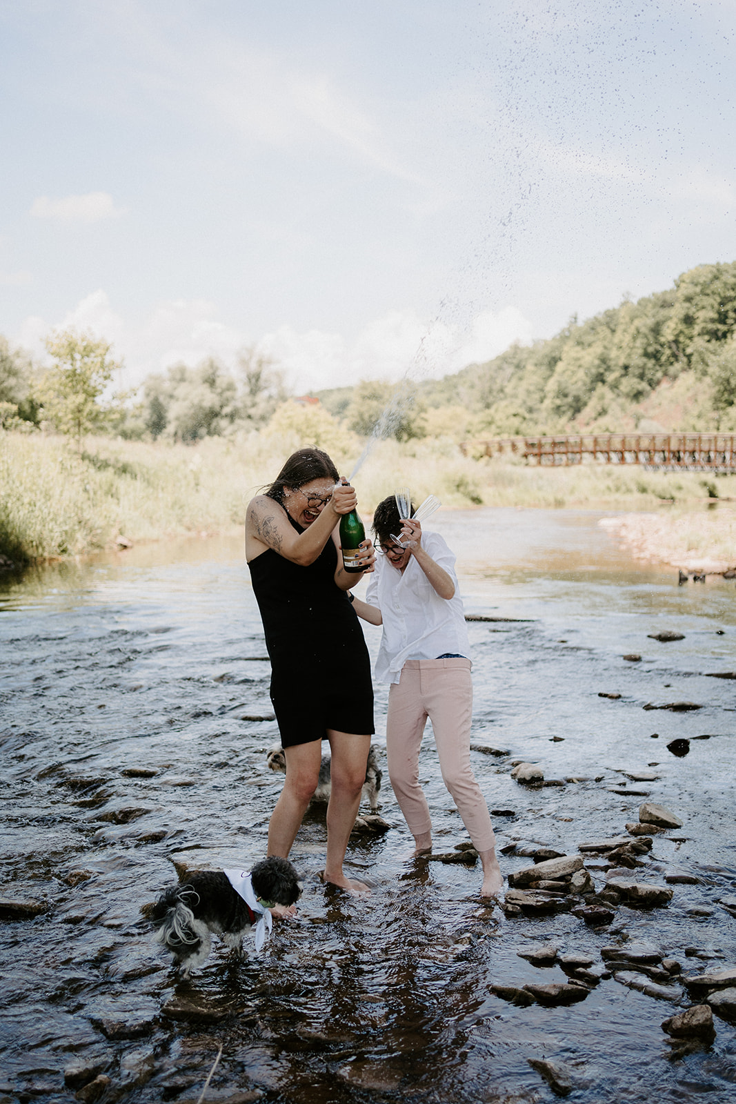 Two people standing on rocks in water while one opens champagne. 