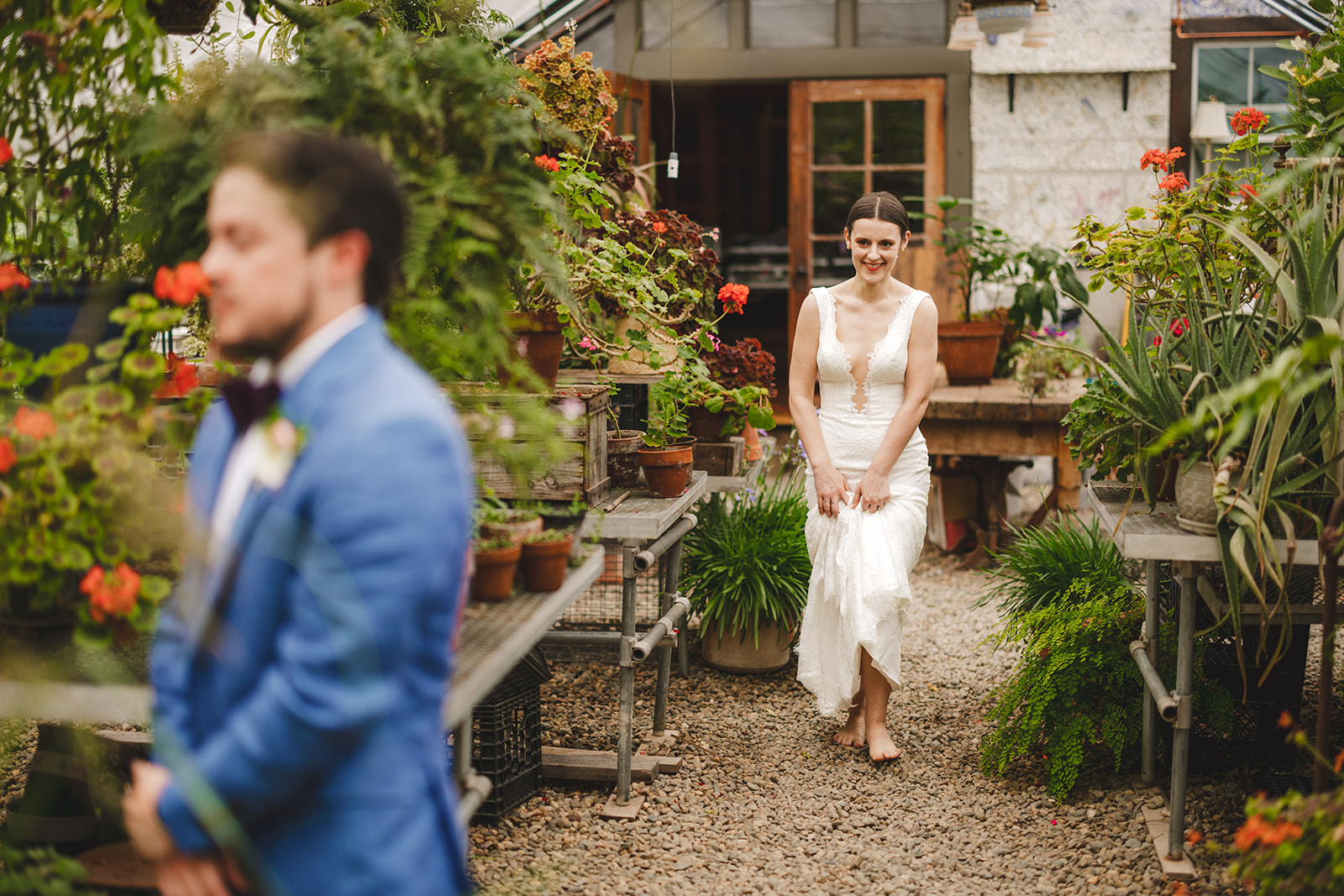 a barefoot bride wearing a white dress approaches her groom in a blue suit inside a greenhouse 