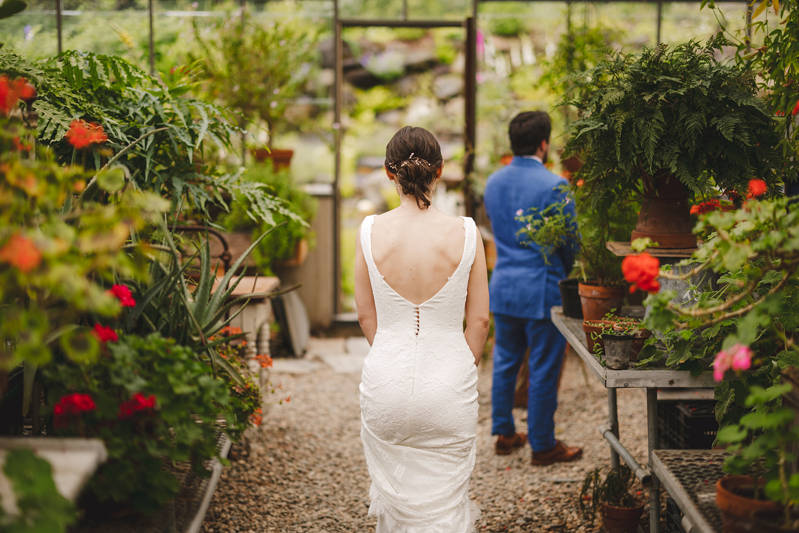 a barefoot bride wearing a white dress approaches her groom in a blue suit inside a greenhouse 