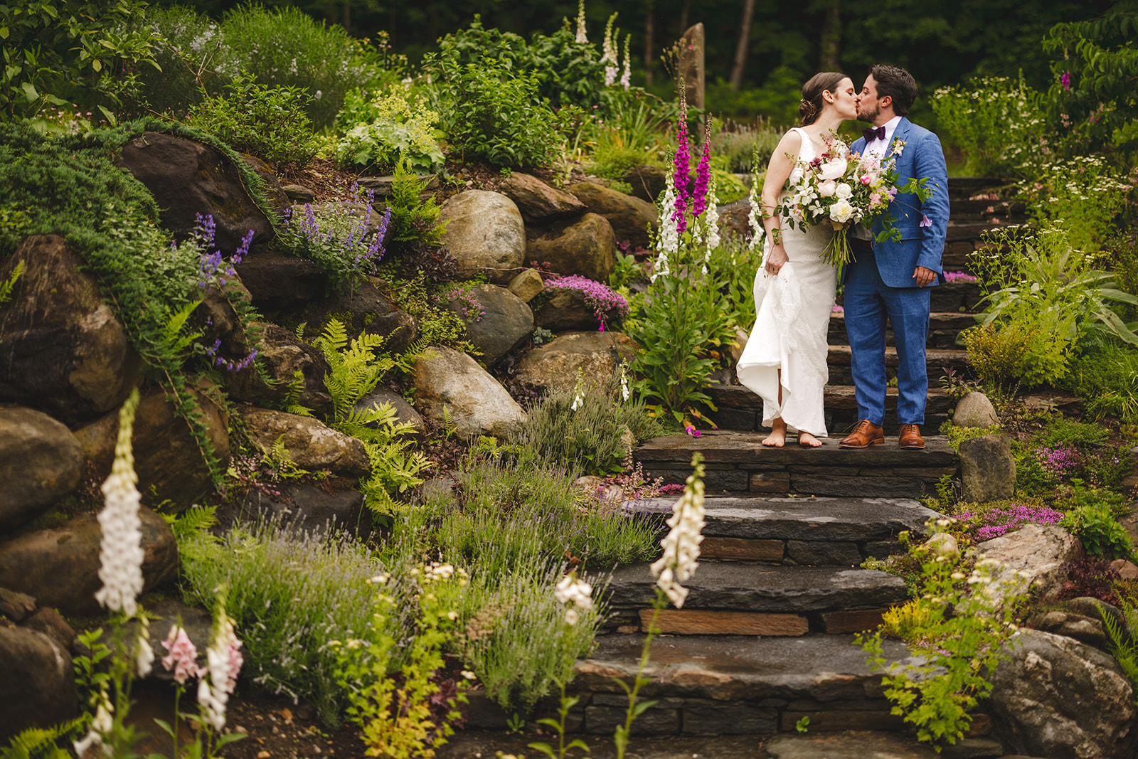 a barefoot bride in a white dress and her groom in a blue suit pose kiss on stone steps in a garden  