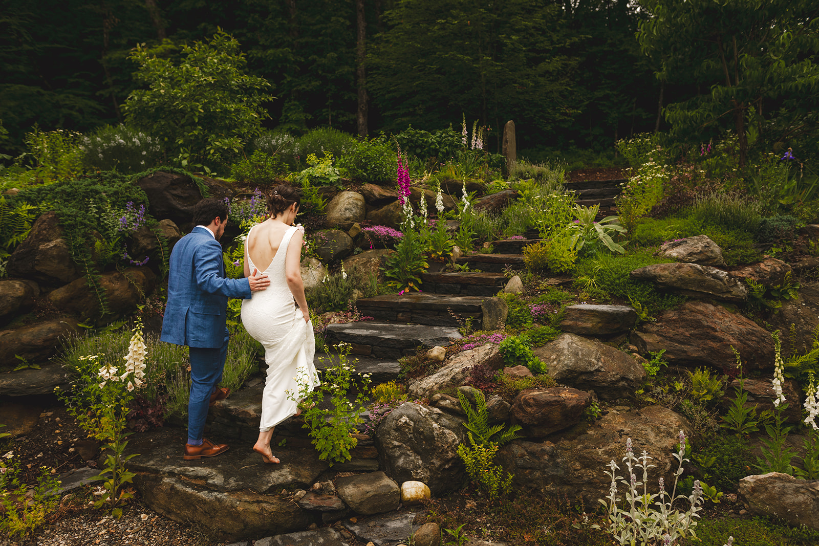 a barefoot bride in a white dress and her groom in a blue suit pose walk up stone steps in a garden  