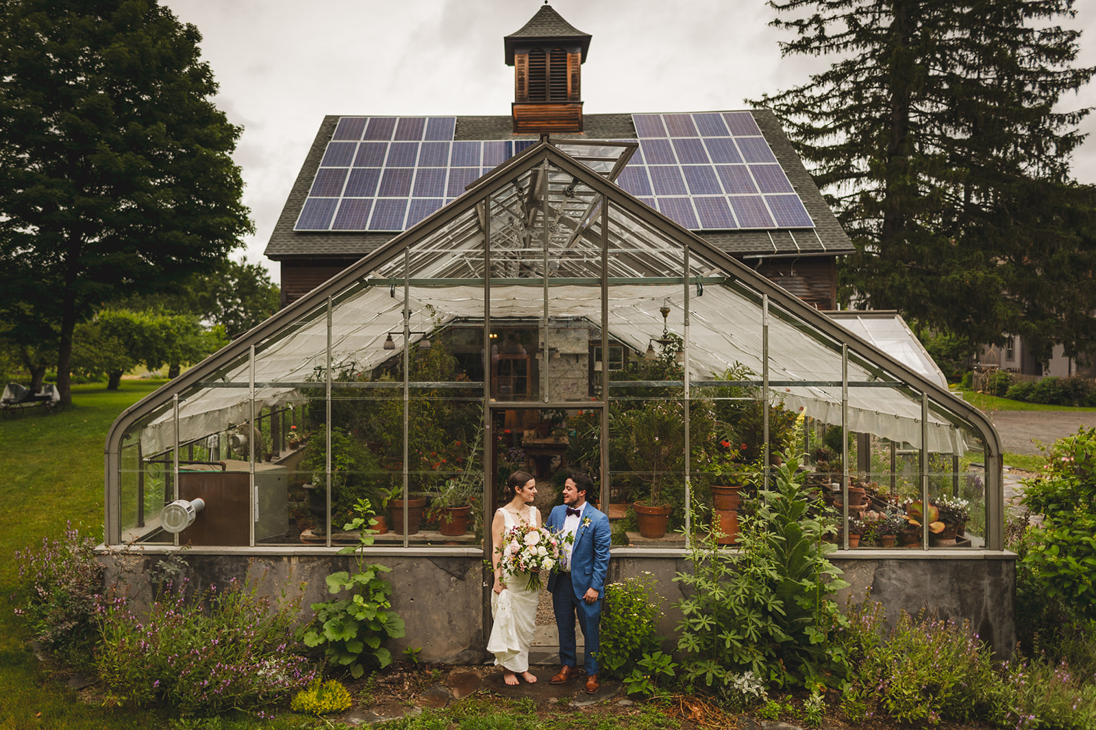 a bride in a white dress and her groom in a blue suit pose in the doorway of a greenhouse at Gloriosa & Co. in Ashfield