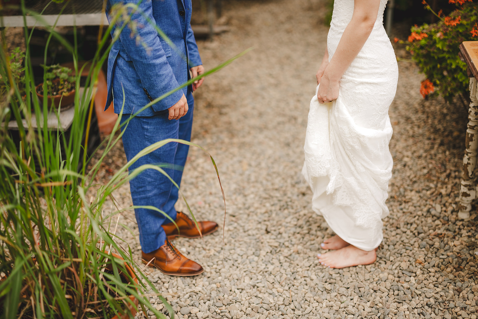 a close up on the legs and feet of a barefoot bride and her groom in his blue suit and brown shoes standing on pebbles 