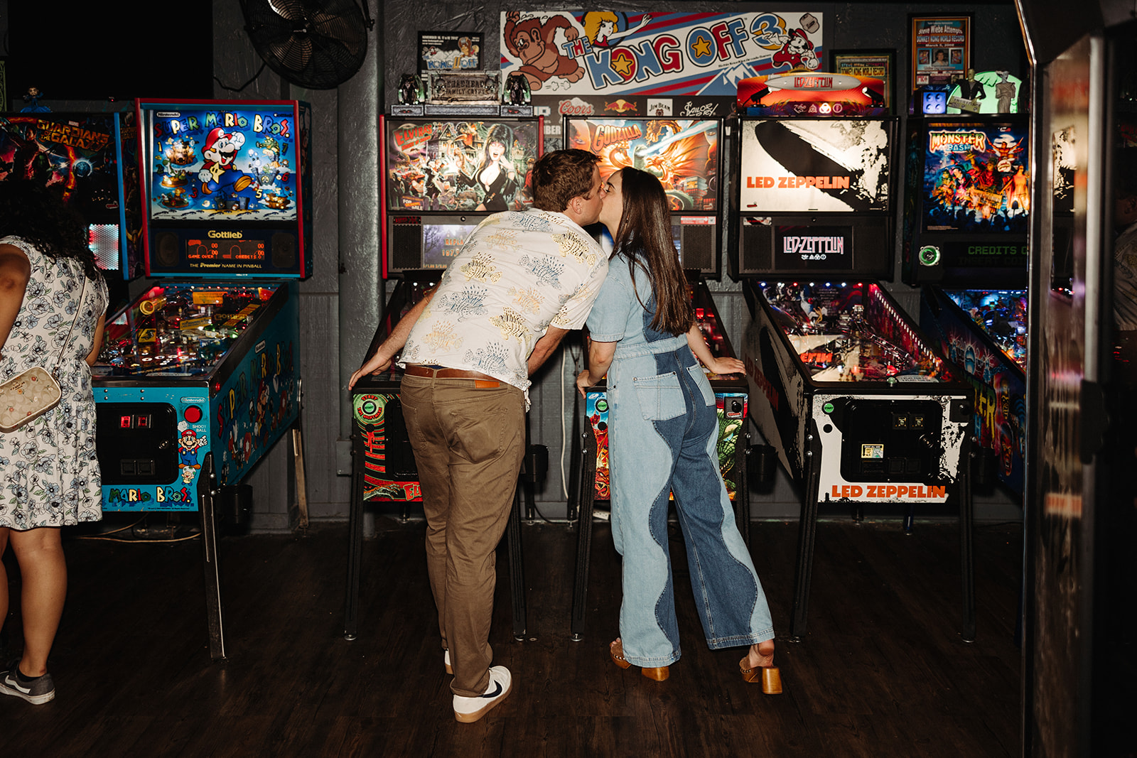 Couple kissing in front of a pinball machine with their backs facing the camera