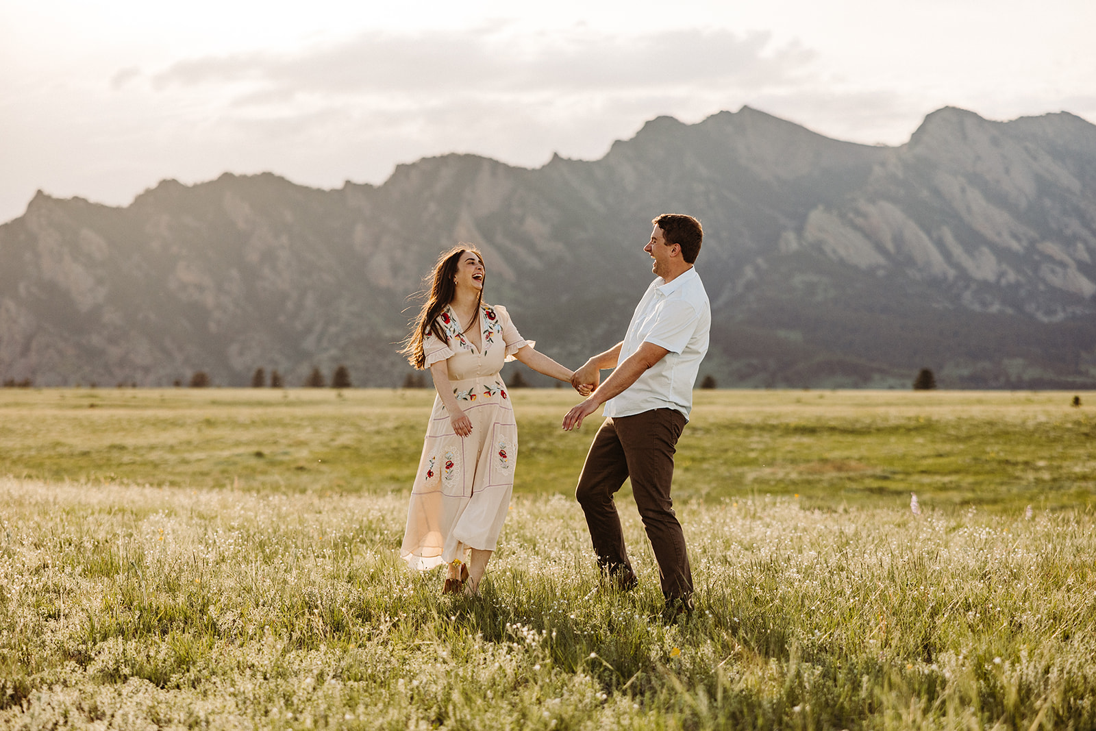 The sun shines on a couple while holding hands and beginning to dance in an open field before a mountain range