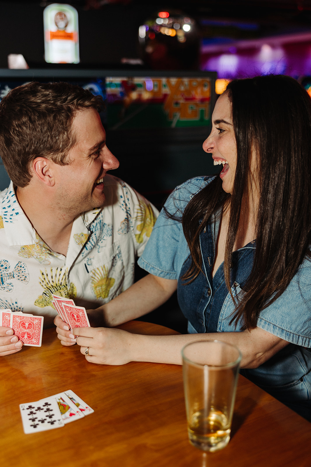 Couple laughing while they play cards and enjoy drinks at their arcade engagement photo shoot