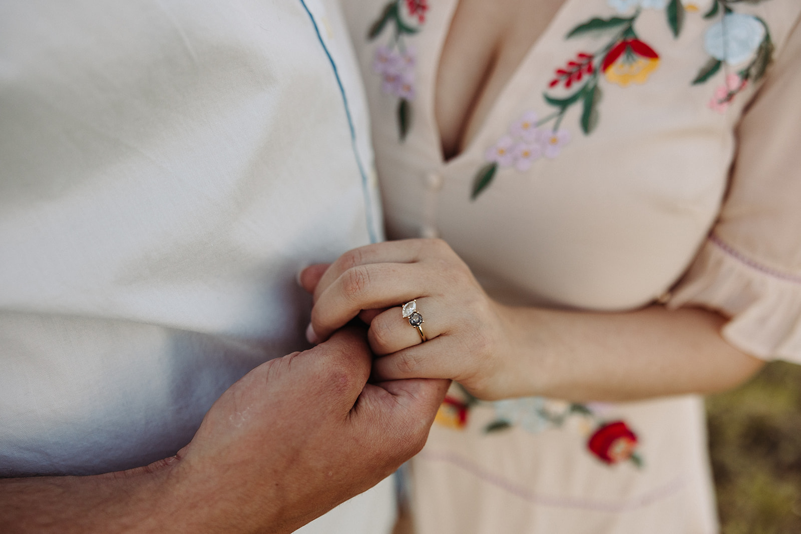 A close-up shot of engaged couple holding hands, the woman's engagement ring, and her embroidered light pink dress