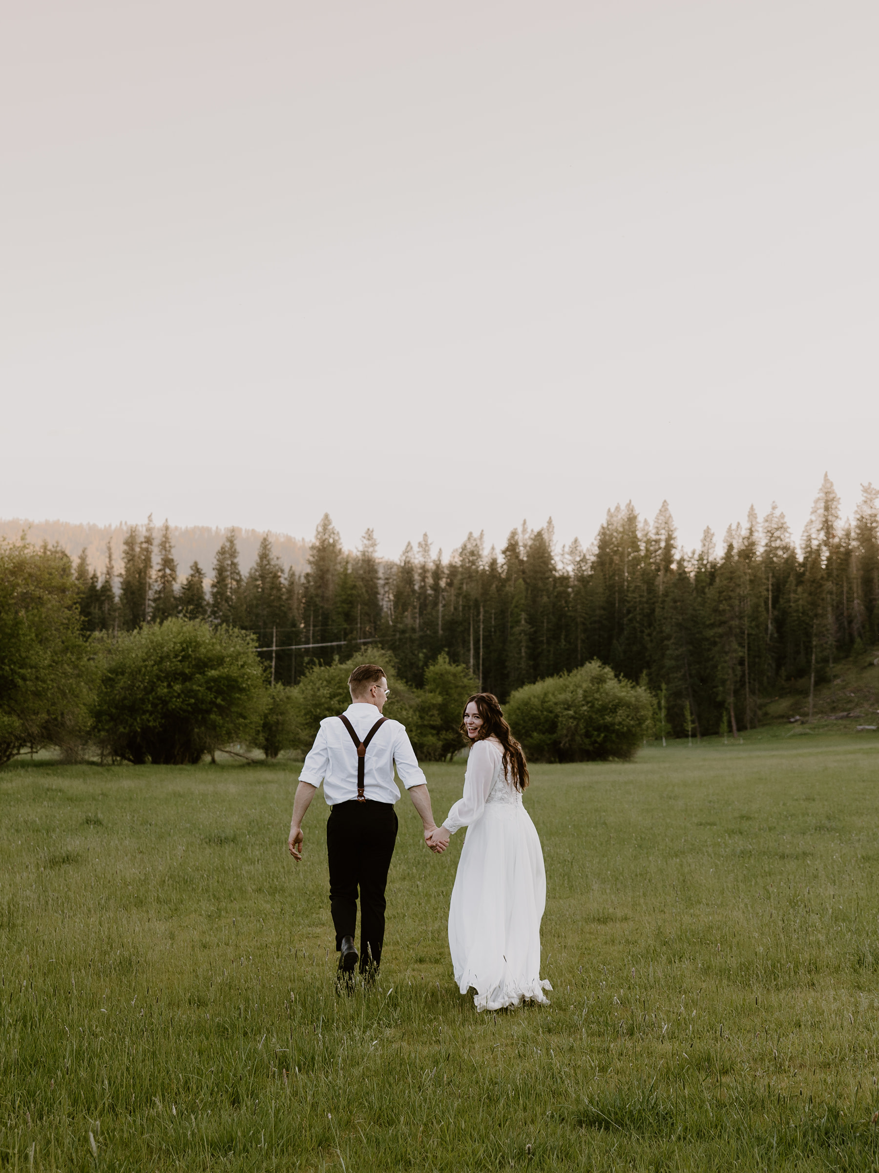 a man and woman in wedding outfits walking in a field, the bride is looking back smiling