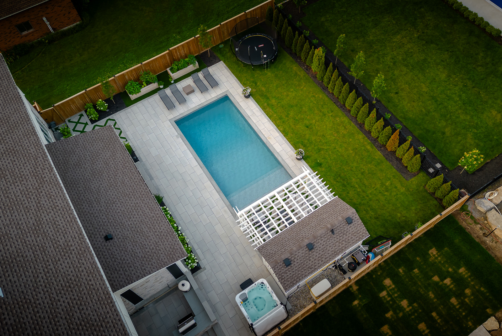 Inground pool with lawn chairs on top of patio stones and a garden along the fence.
