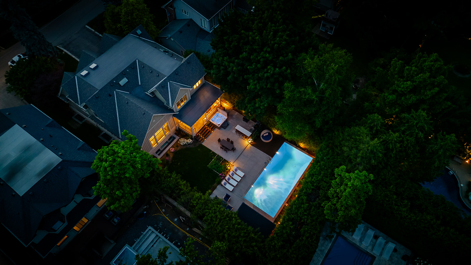 A drone shot of the backyard with the lights on in the pool and inside the house.