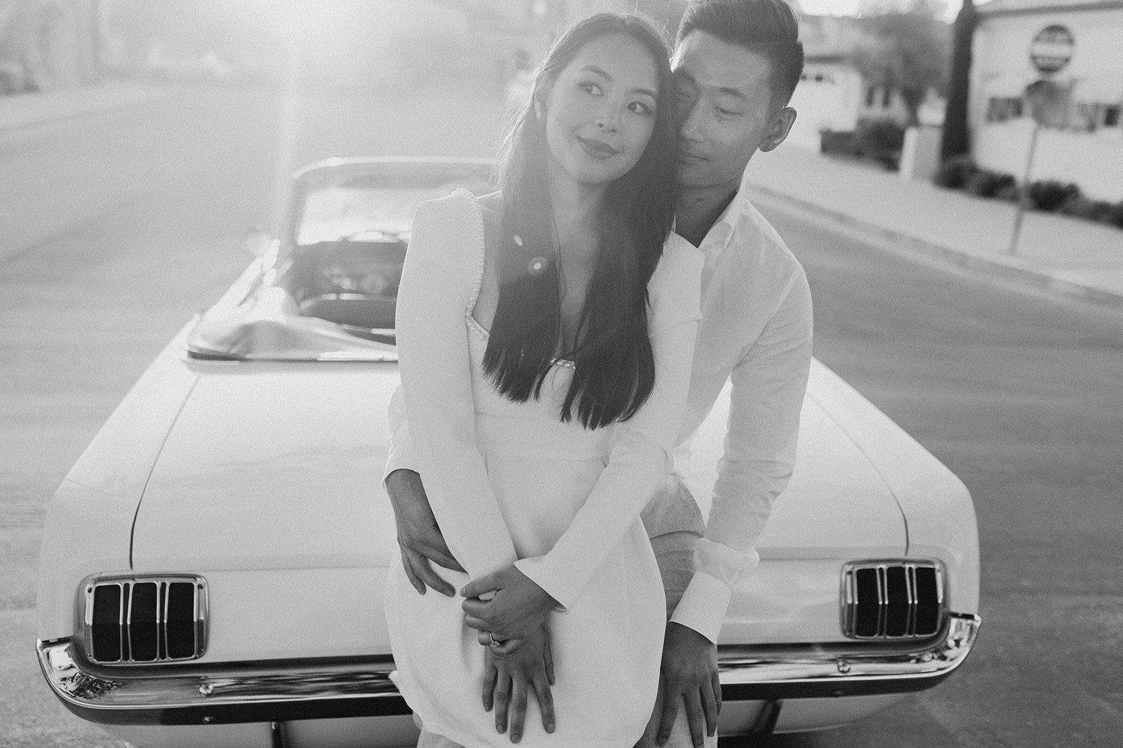 black and white photo of couple leaning against hood of a mustang car with sun shining behind 