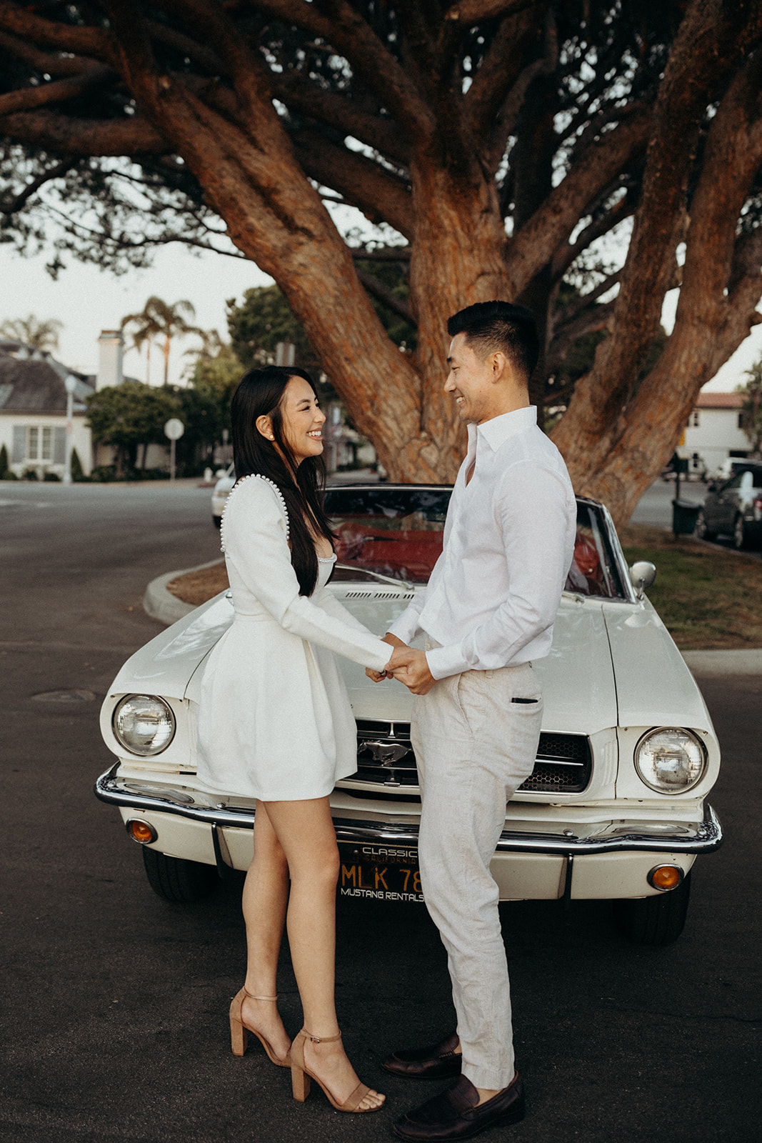 couple dressed in white standing in front of white mustang holding hands