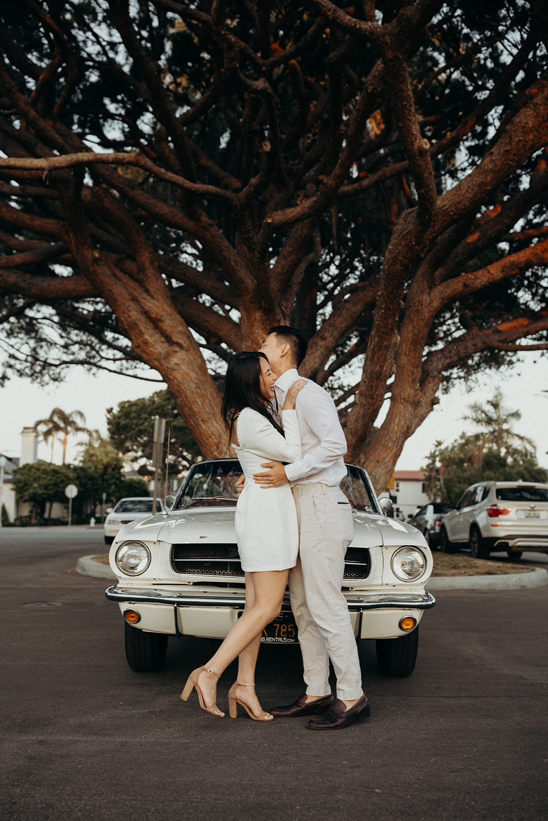couple dressed in white standing in front of white mustang laughing