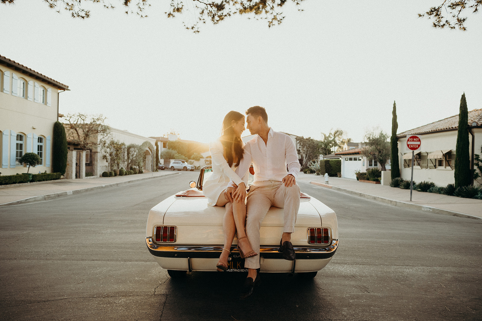 couple leaning forehead to forehead sitting on the hood of a  white vintage mustang while sun sets 