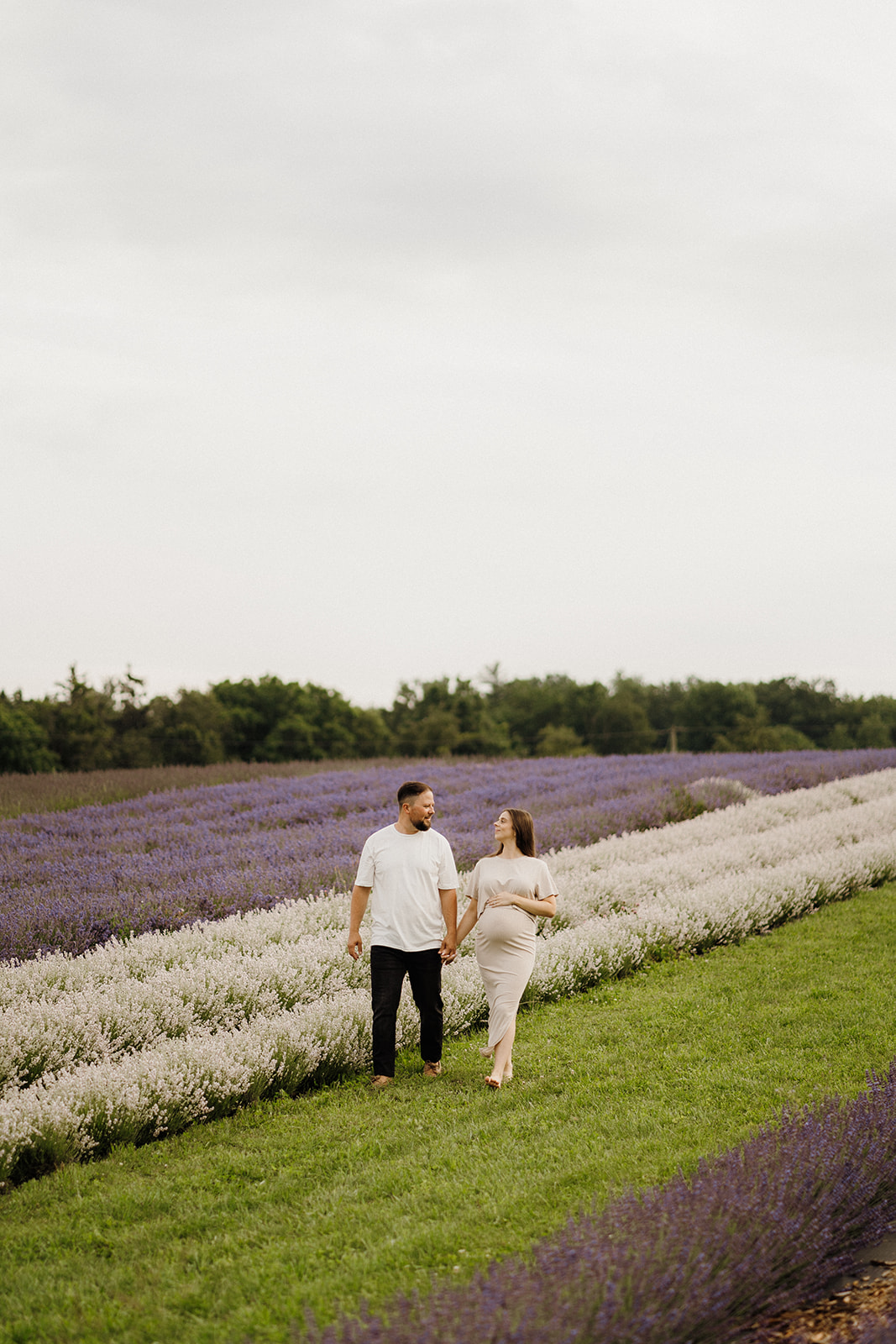 Man and woman hold hands while walking on the grass looking at each other by the lavender fields.