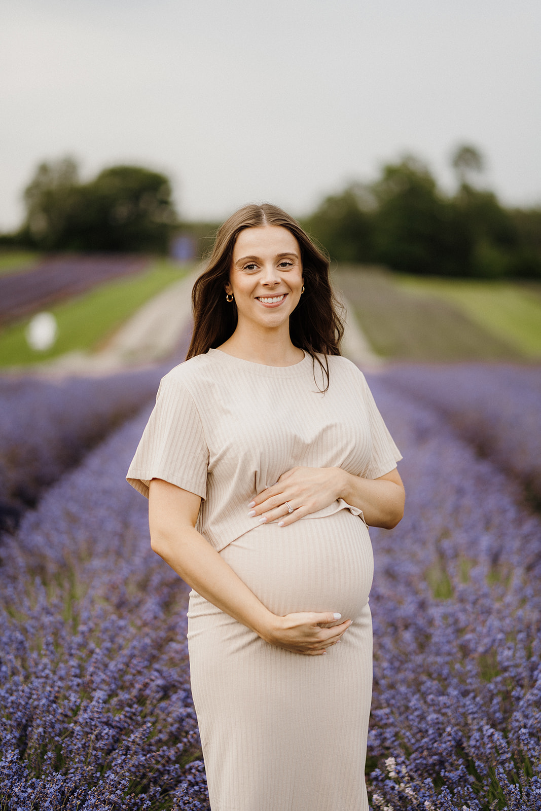 Woman stands between the lavender while resting her hands on both top and bottom of her stomach.