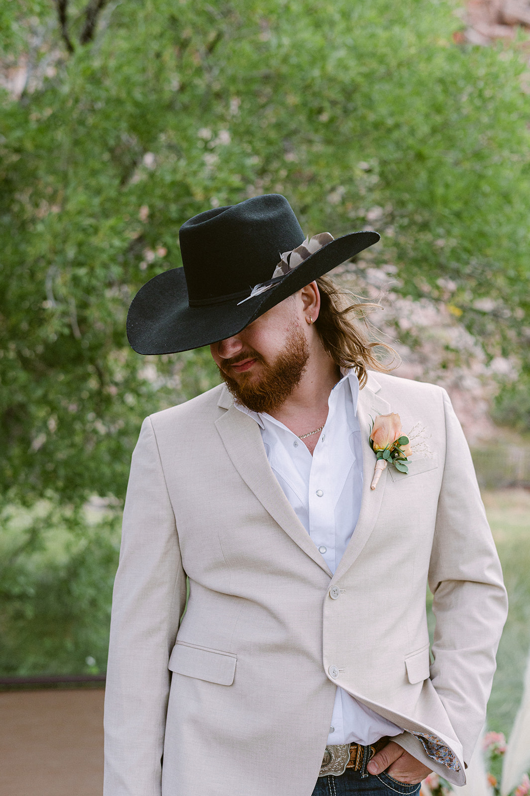 groom posing with black cowboy hat and tan jacket with lush greenery at red rock canyon