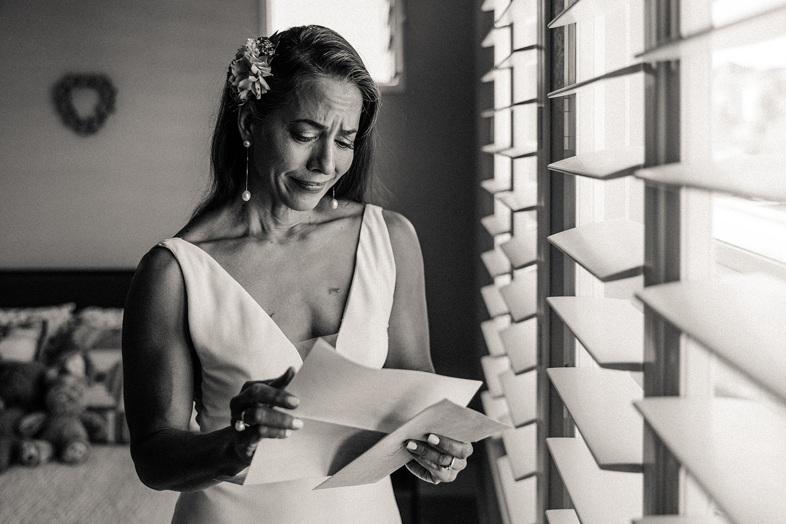 The bride tearfully reading a letter from her groom while she stands in front of a large window, showered in light.