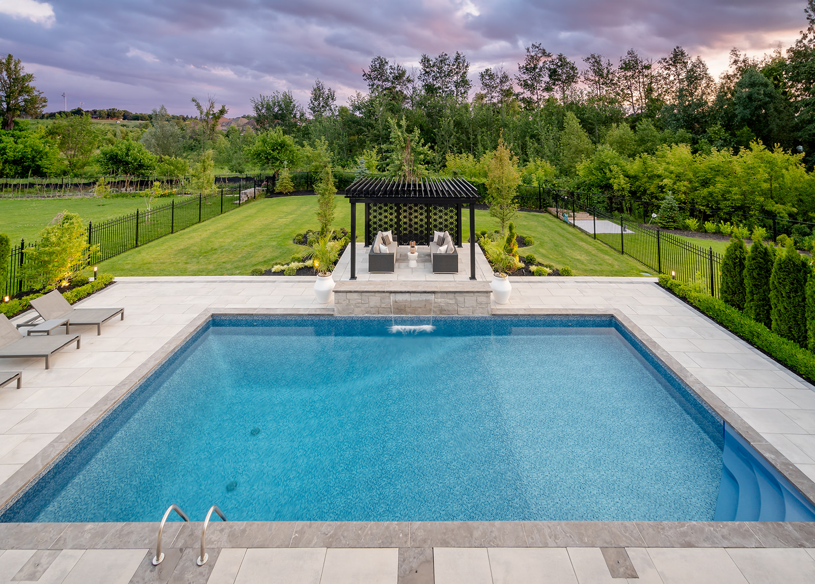 An inground pool with lounge chairs on the left and a metal gazebo near the back with the waterfall on.