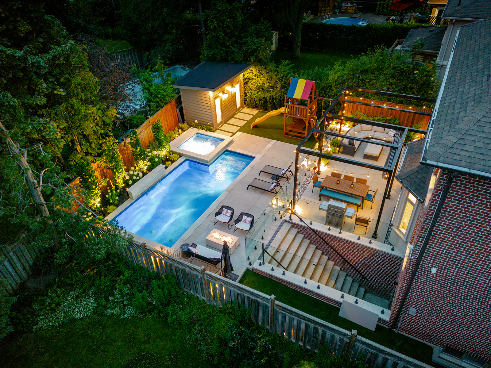 A large backyard, lights on, with an inground pool and furniture.