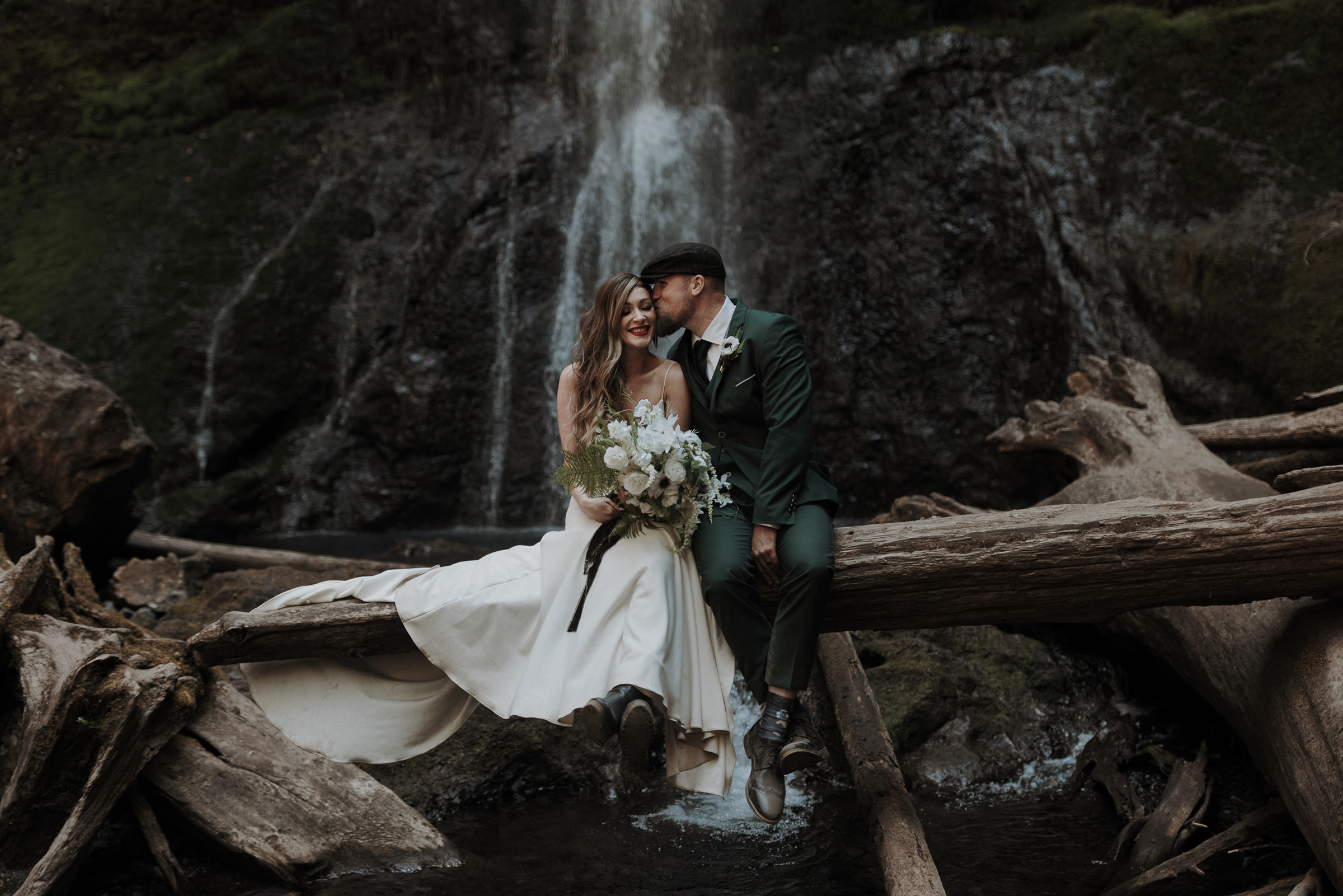 An eloping couple in a white wedding dress and an emerald green suit sitting on a log at Marymere Falls. 