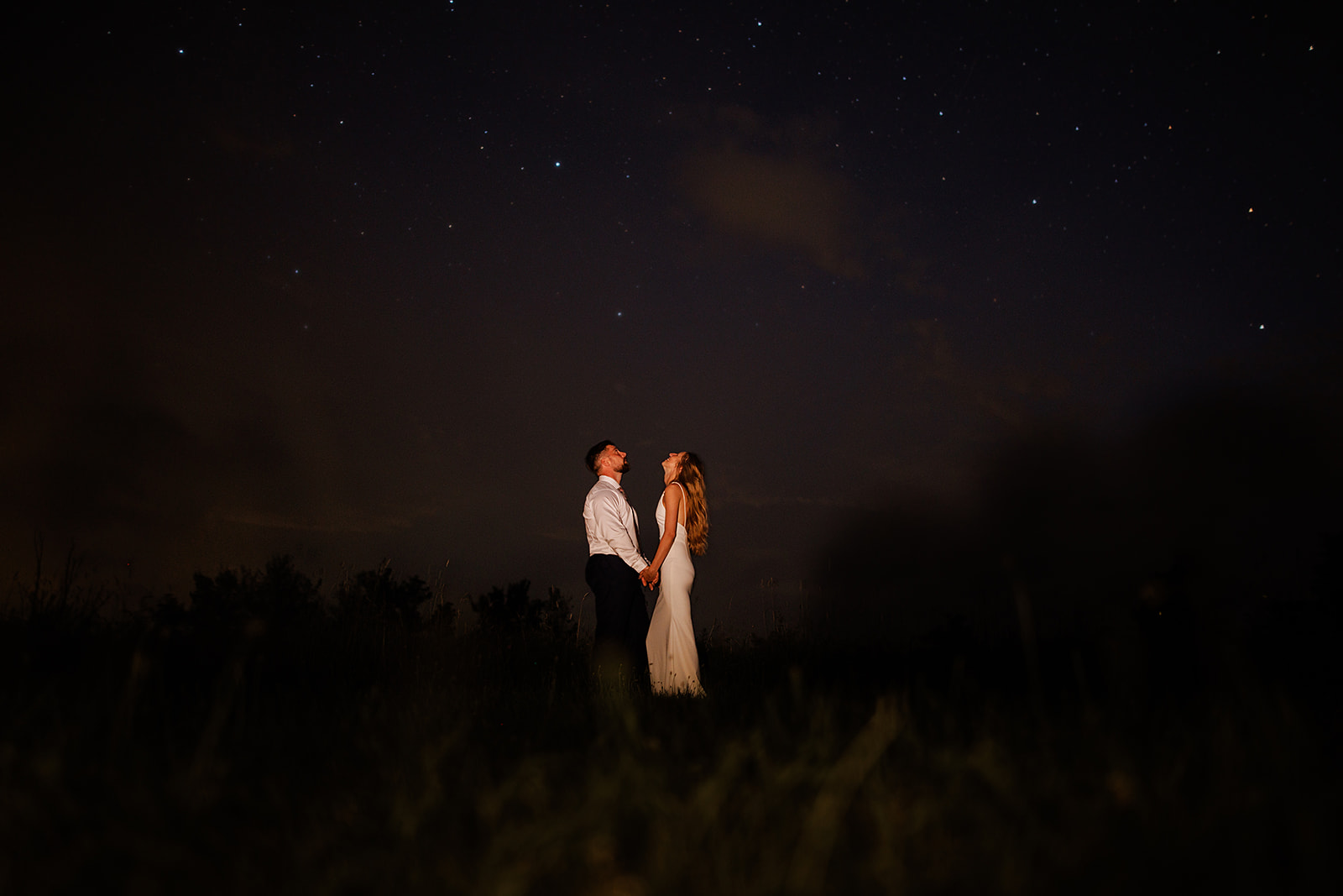 Bride and Groom stand in Catskill field and look up at star filled night sky for a creative portrait on wedding night.