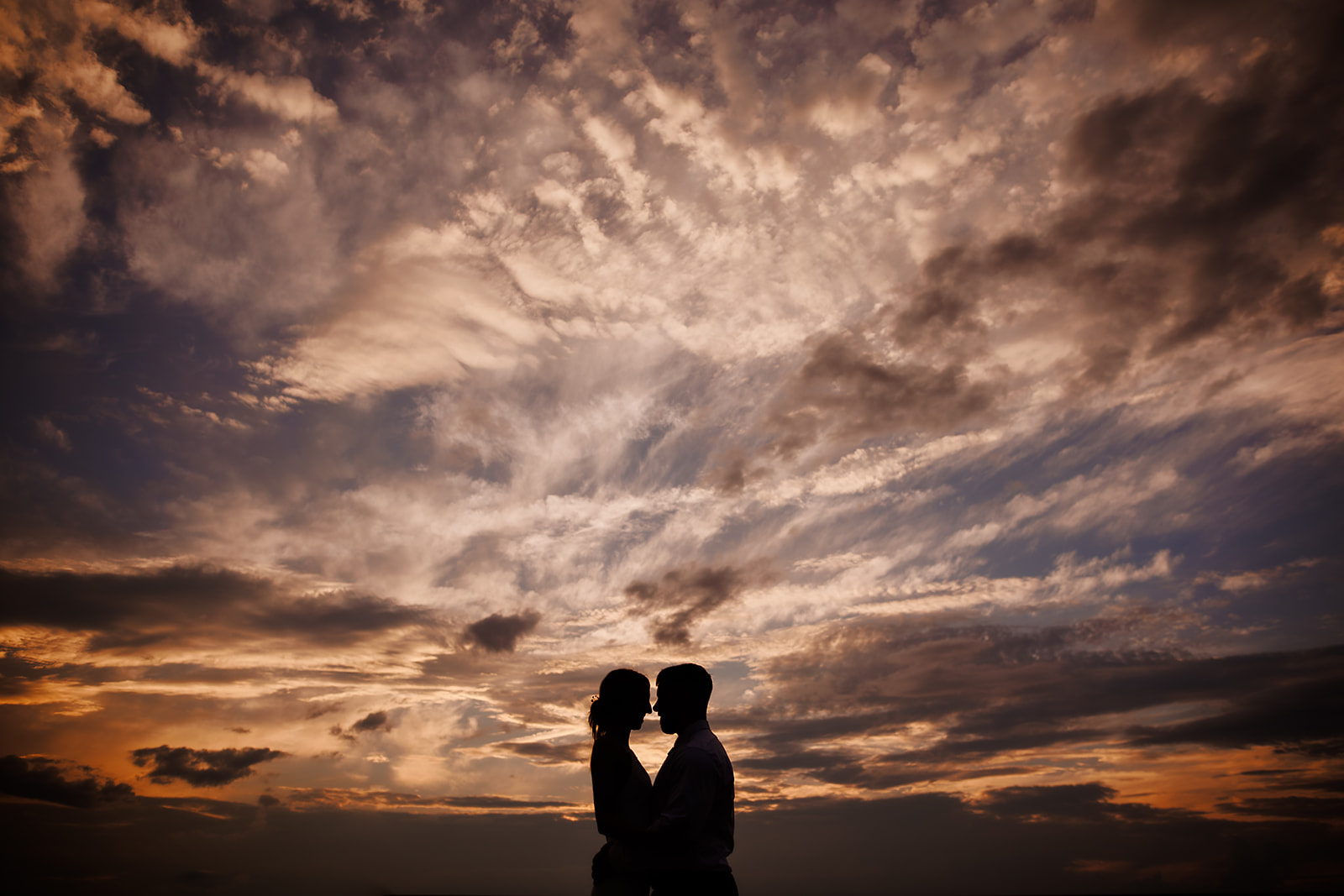Silhouette of just married couple against an amazing sunset sky with detailed orange and pink clouds.