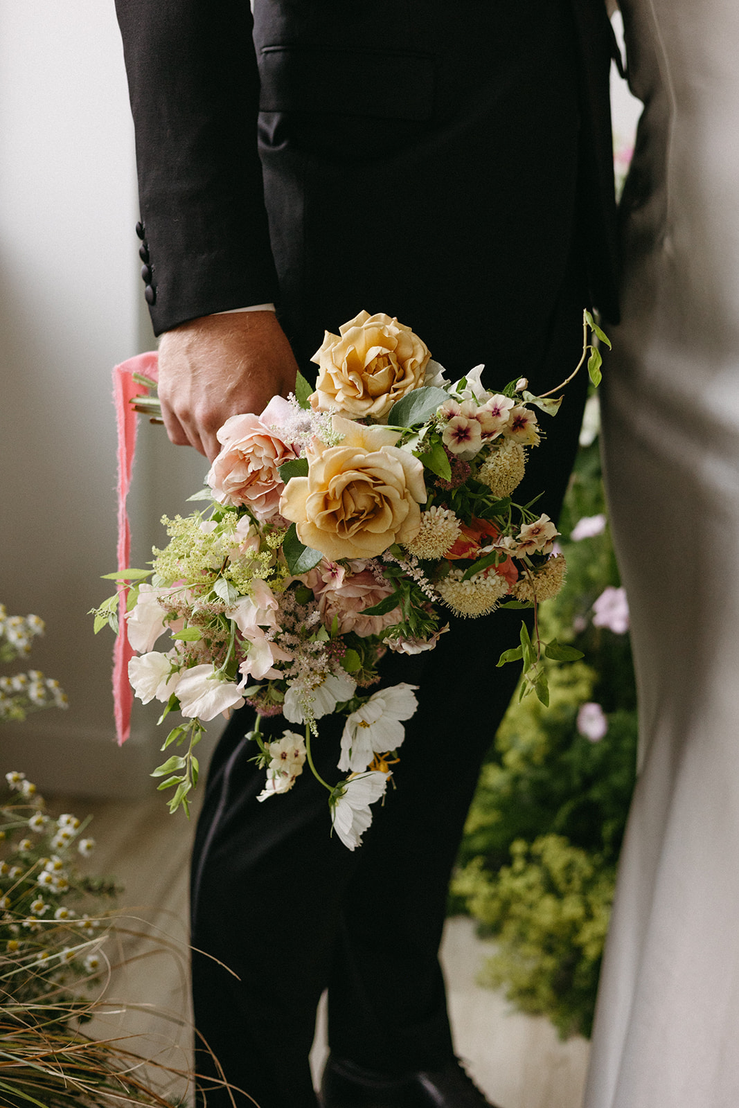 A garden party wedding groom in a fitted black suit holds a soft-colored bridal bouquet tied off with a pink ribbon. 