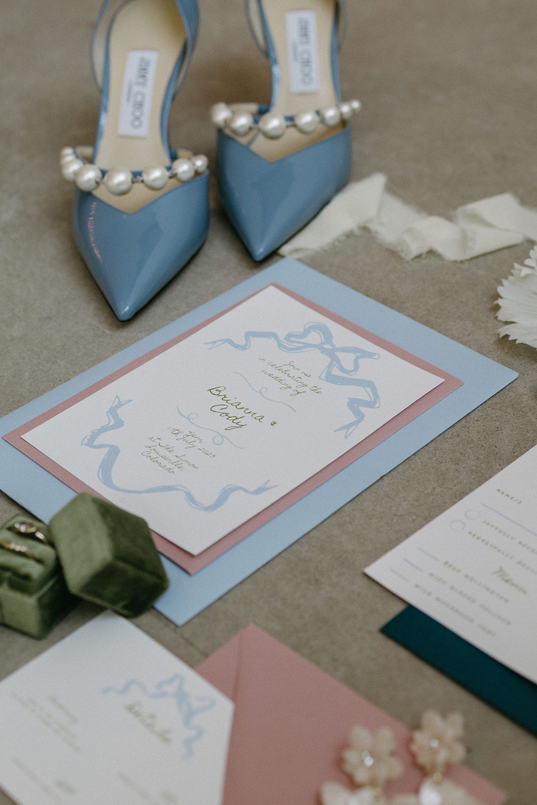 A garden party wedding flat lay featuring a baby blue, dusty rose invitation suite, Jimmy Choo heels, and green ring box