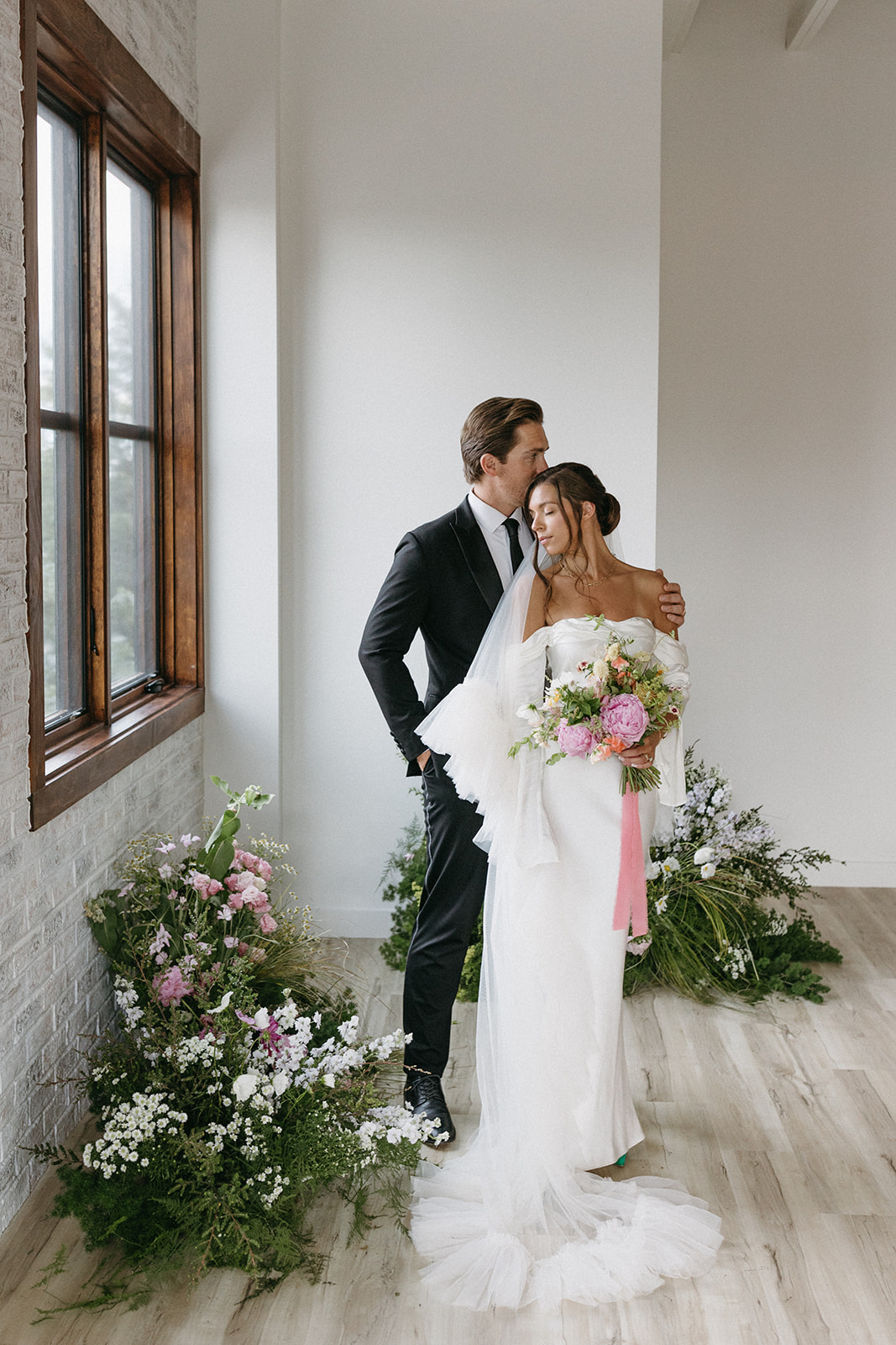 A groom in a designer suit kisses a bride on the head as she faces a large window in a room filled with natural light. 
