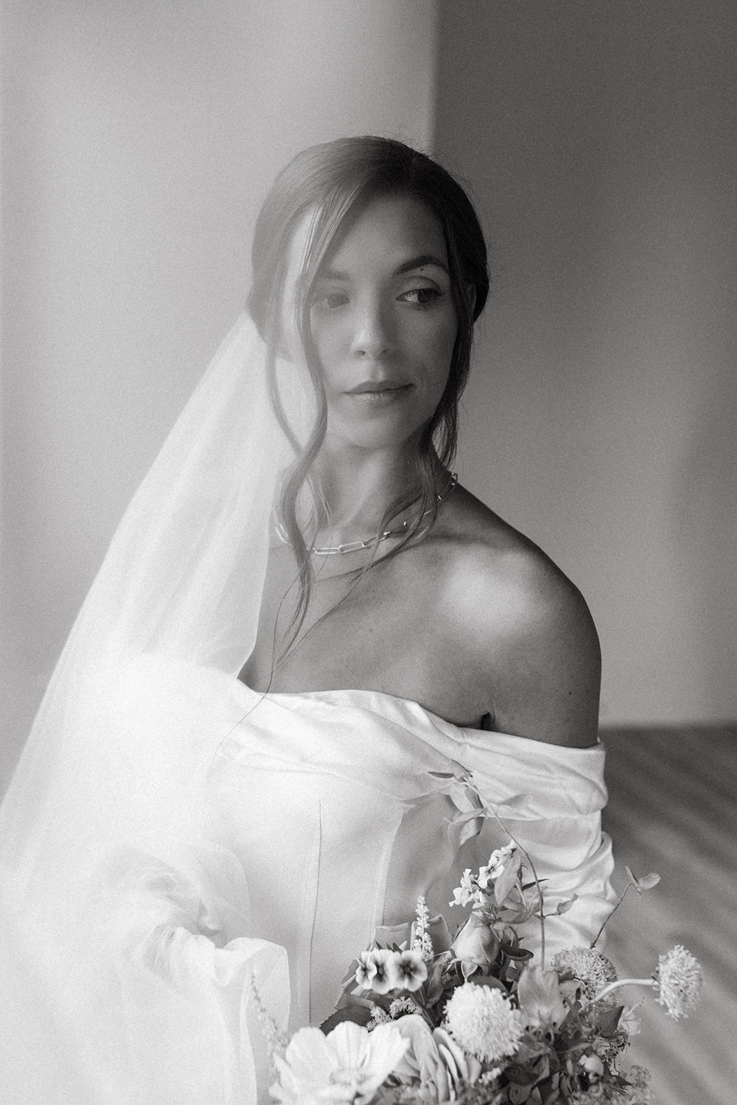 A bride wearing an elegant updo wedding hairstyle and long tulle bridal veil looks into the distance, holding a bouquet.