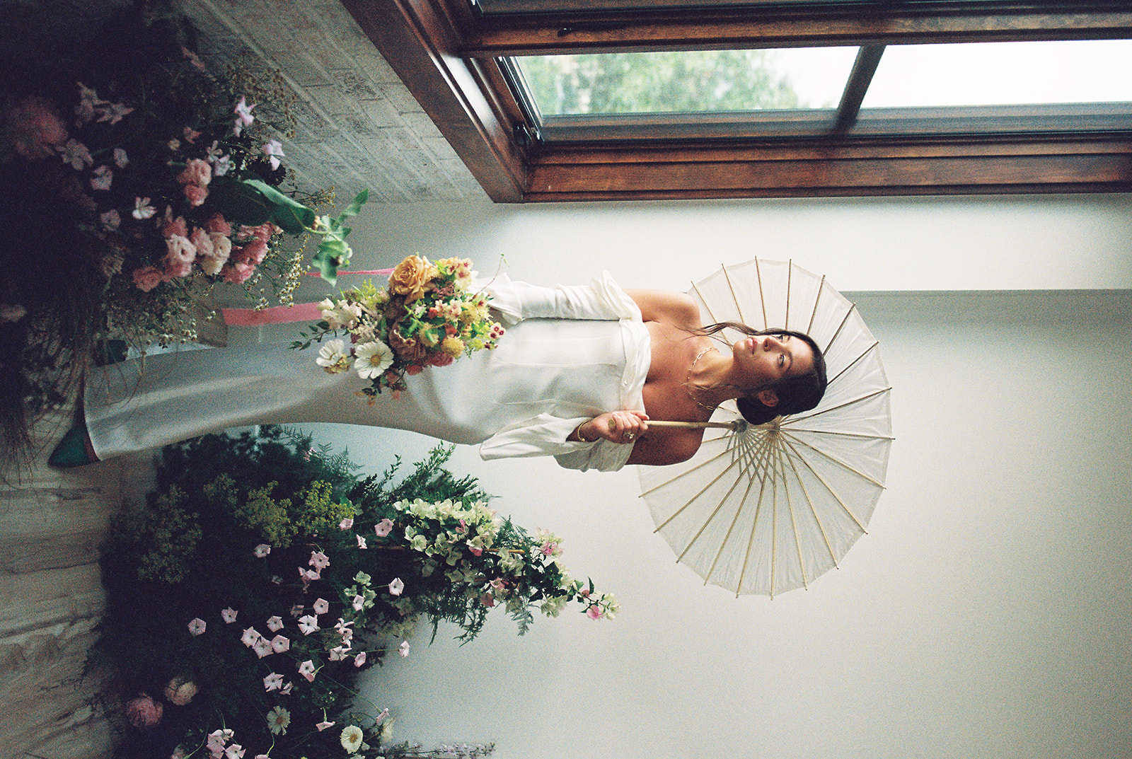 An elegant bride in an off-the-shoulder wedding dress holding a white paper parasol and bright floral bridal bouquet.  