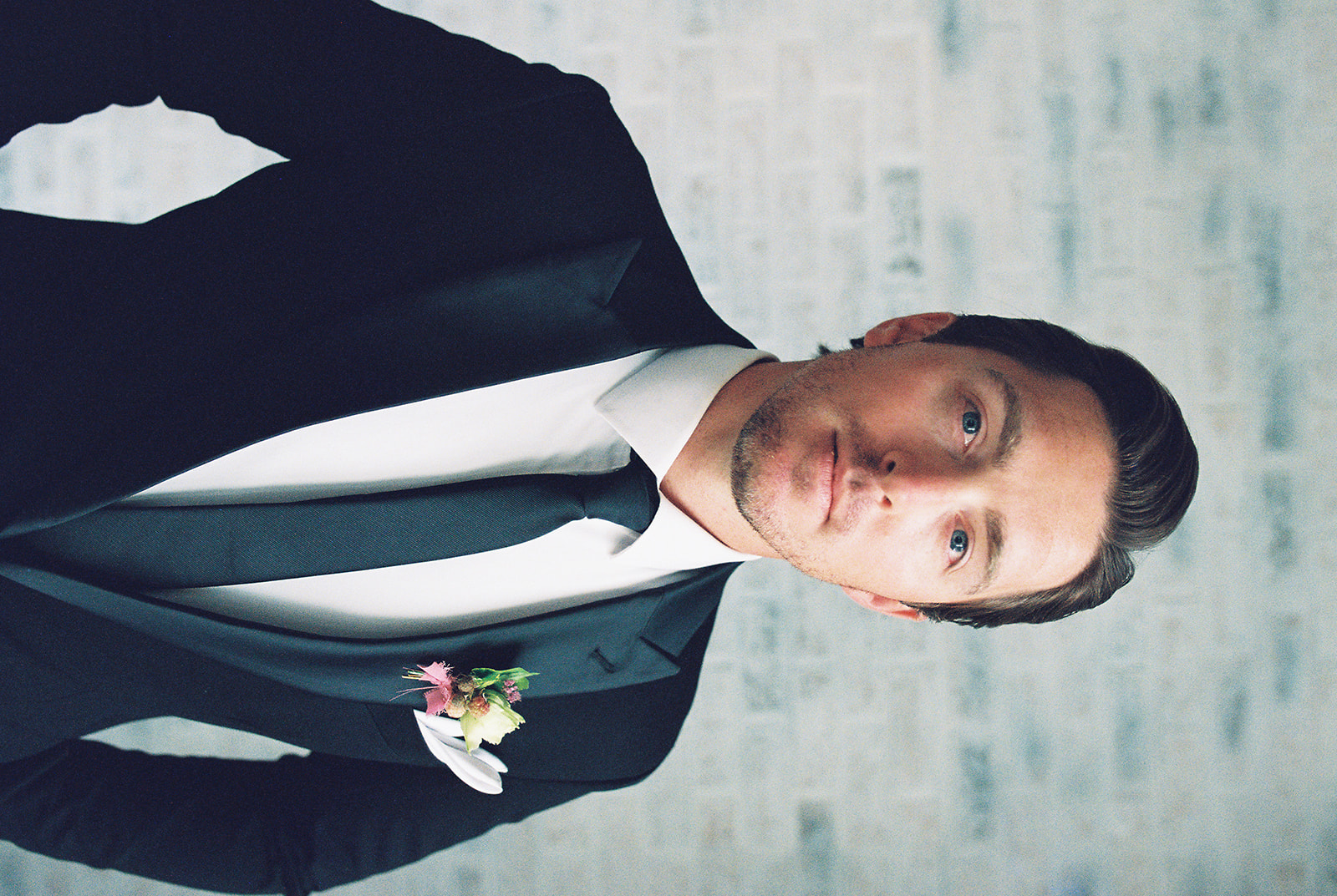A wedding portrait of a groom in a designer black suit featuring a fresh floral boutonniere on his jacket's lapel.