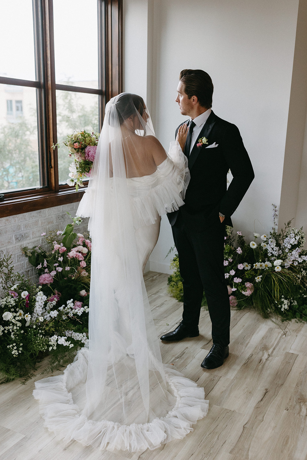 A bride wearing an off-the-shoulder dress and chapel-length bridal veil rests her hand on her groom's shoulder. 