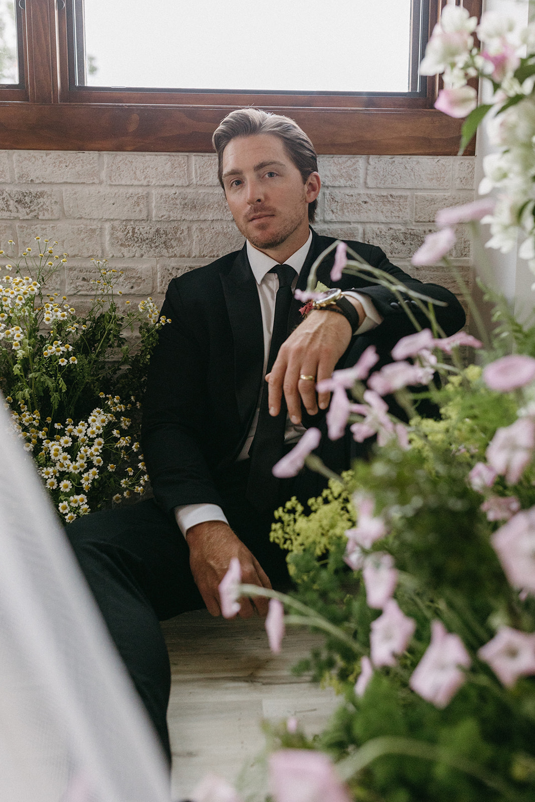 A groom softly smiles while sitting, leaning on a brick wall and surrounded by garden party wedding florals. 