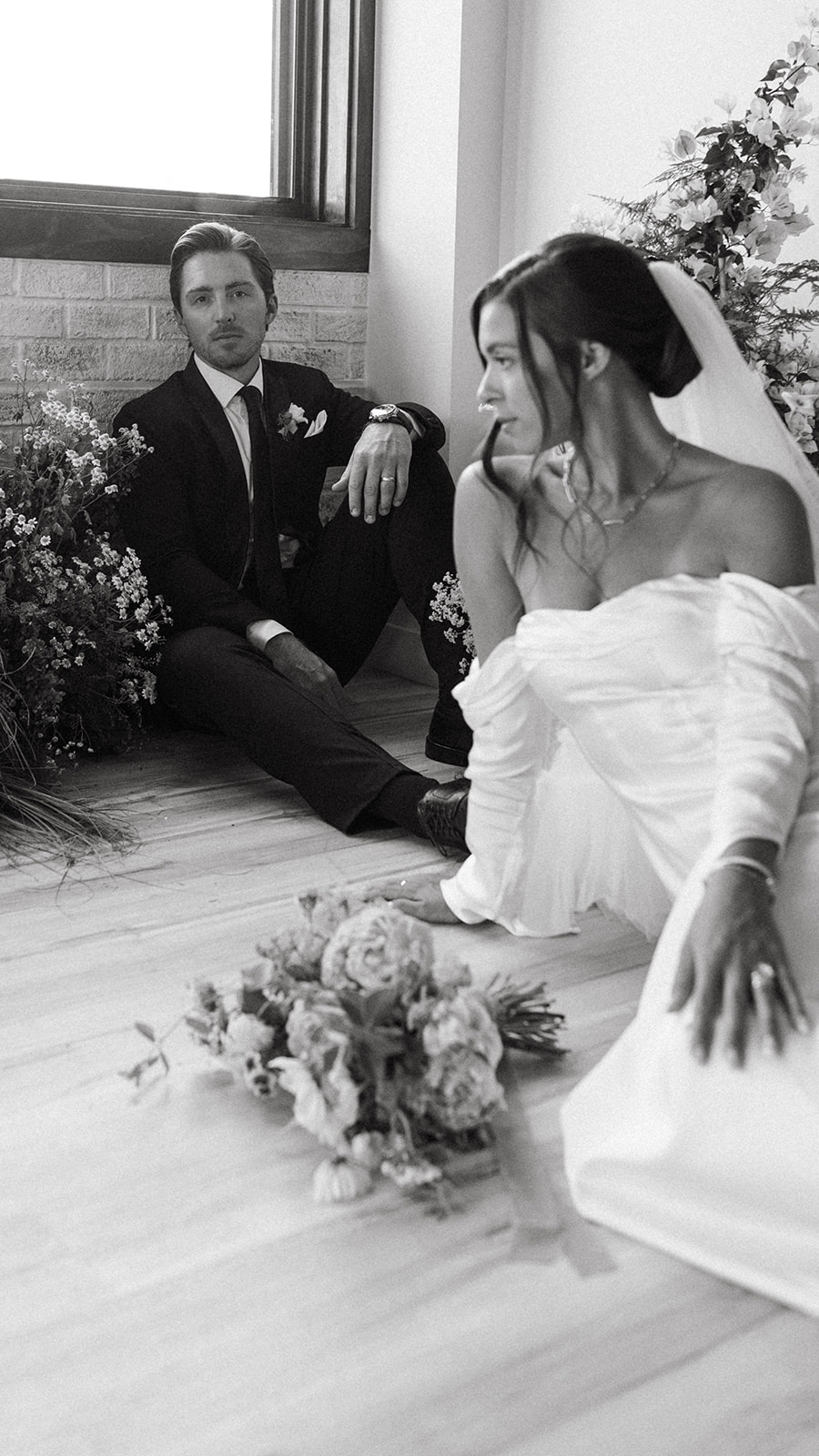 A groom in a designer suit sits on a wood floor with a bride who is wearing an off-the-shoulder wedding dress.  