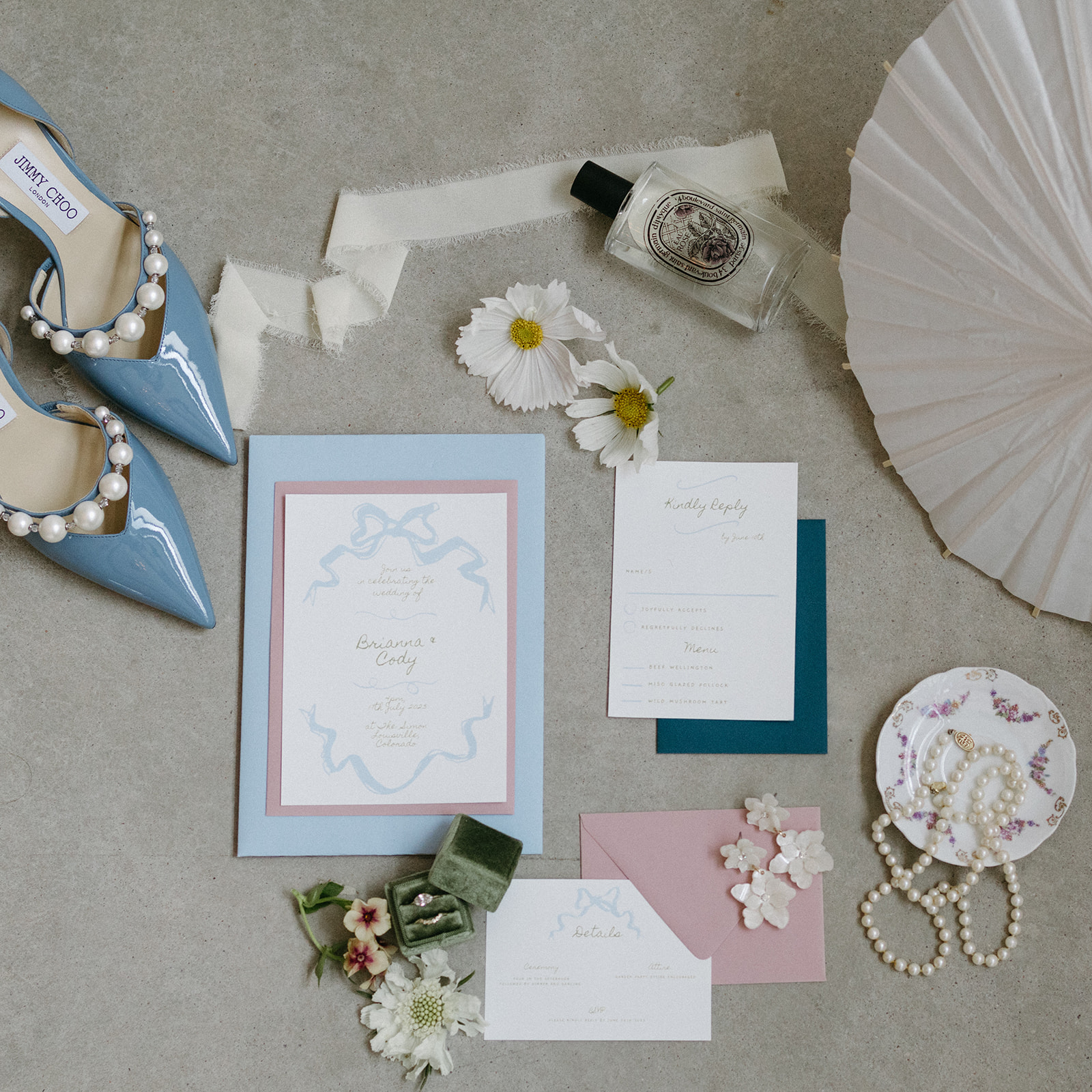 An elegant wedding flat lay featuring a baby blue, dusty rose invitation suite, Jimmy Choo wedding heels, and pearls. 
