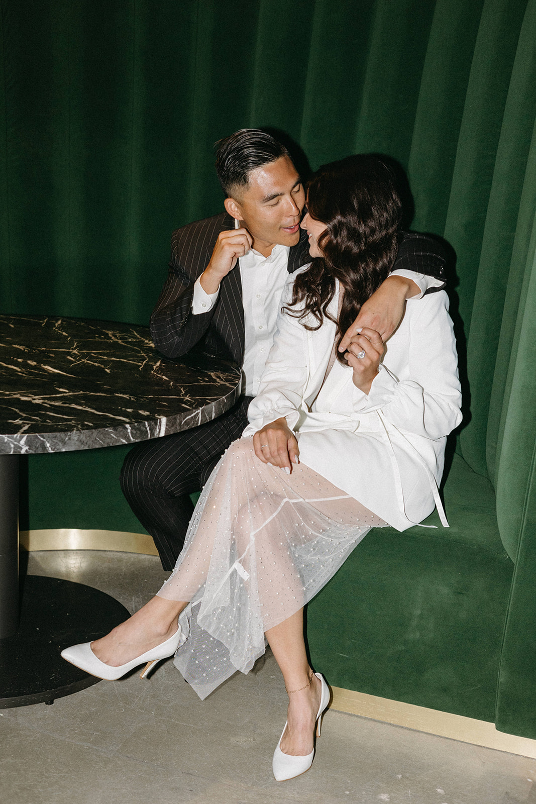 A speakeasy wedding bride and groom are seated in a green velvet booth in timeless wedding attire.