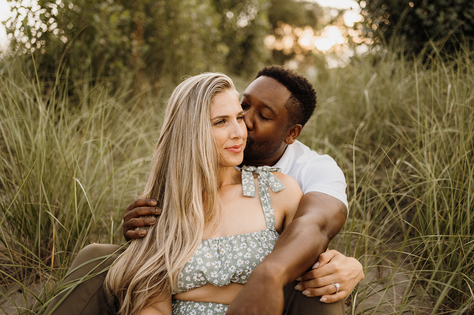 Woman sits in between mans legs on the grass. Man kisses her cheek.