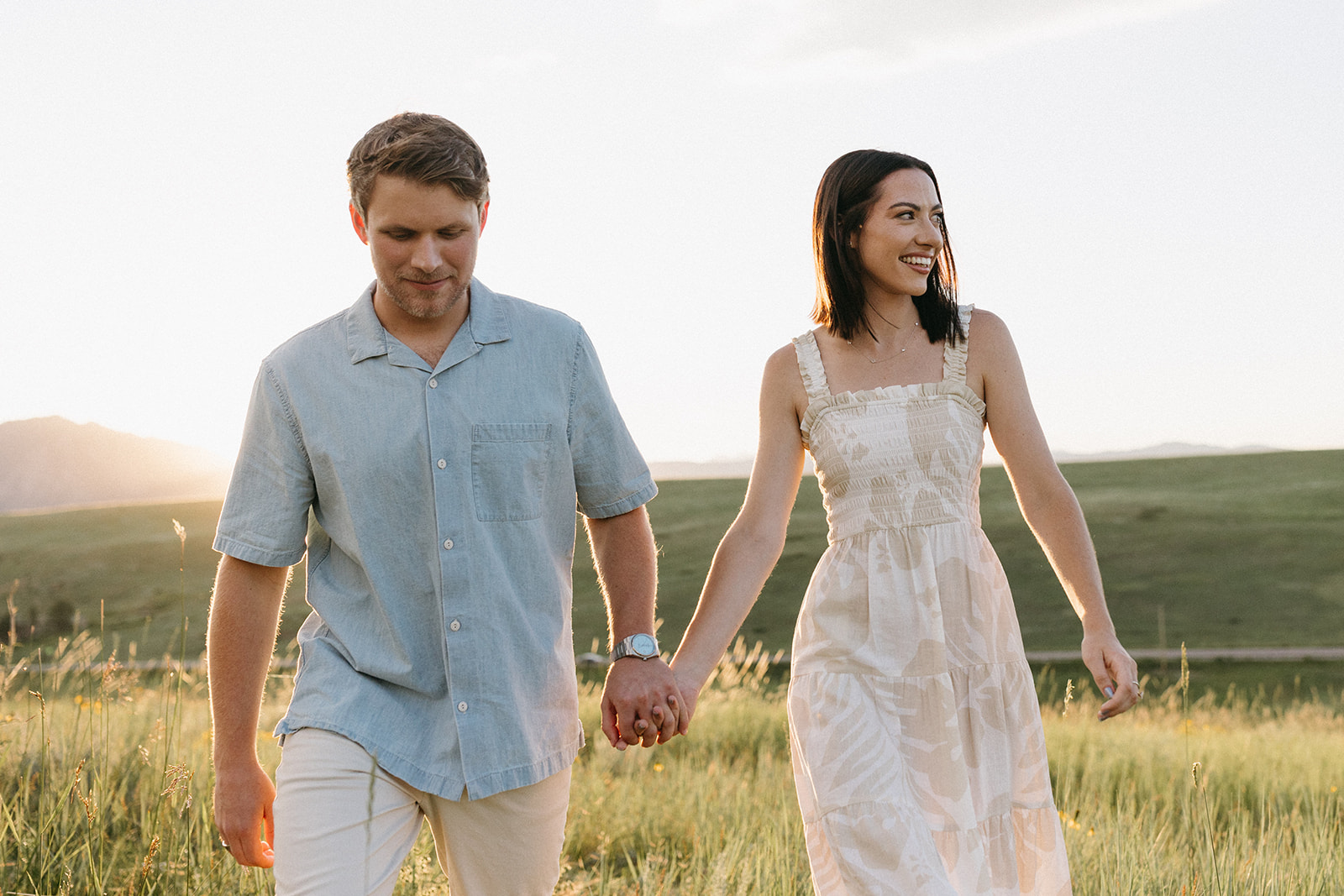 An engaged couple dressed in neutral-toned attire holds hands while walking through a wildflower meadow. 