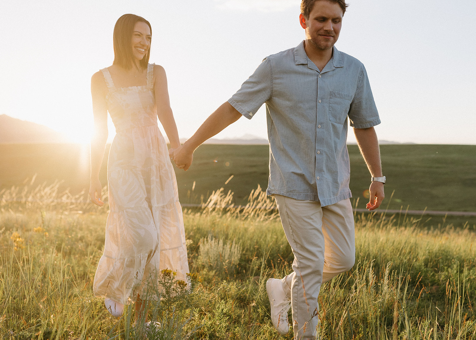 A Boulder, Colorado, Sunset shines on an editorial engagement couple who are holding hands and walking through a field. 