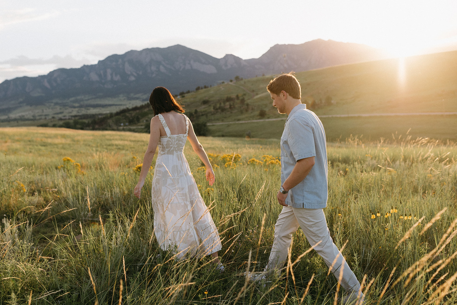 An editorial engagement session couple wearing natural-tone outfits walks through a wildflower meadow near Boulder