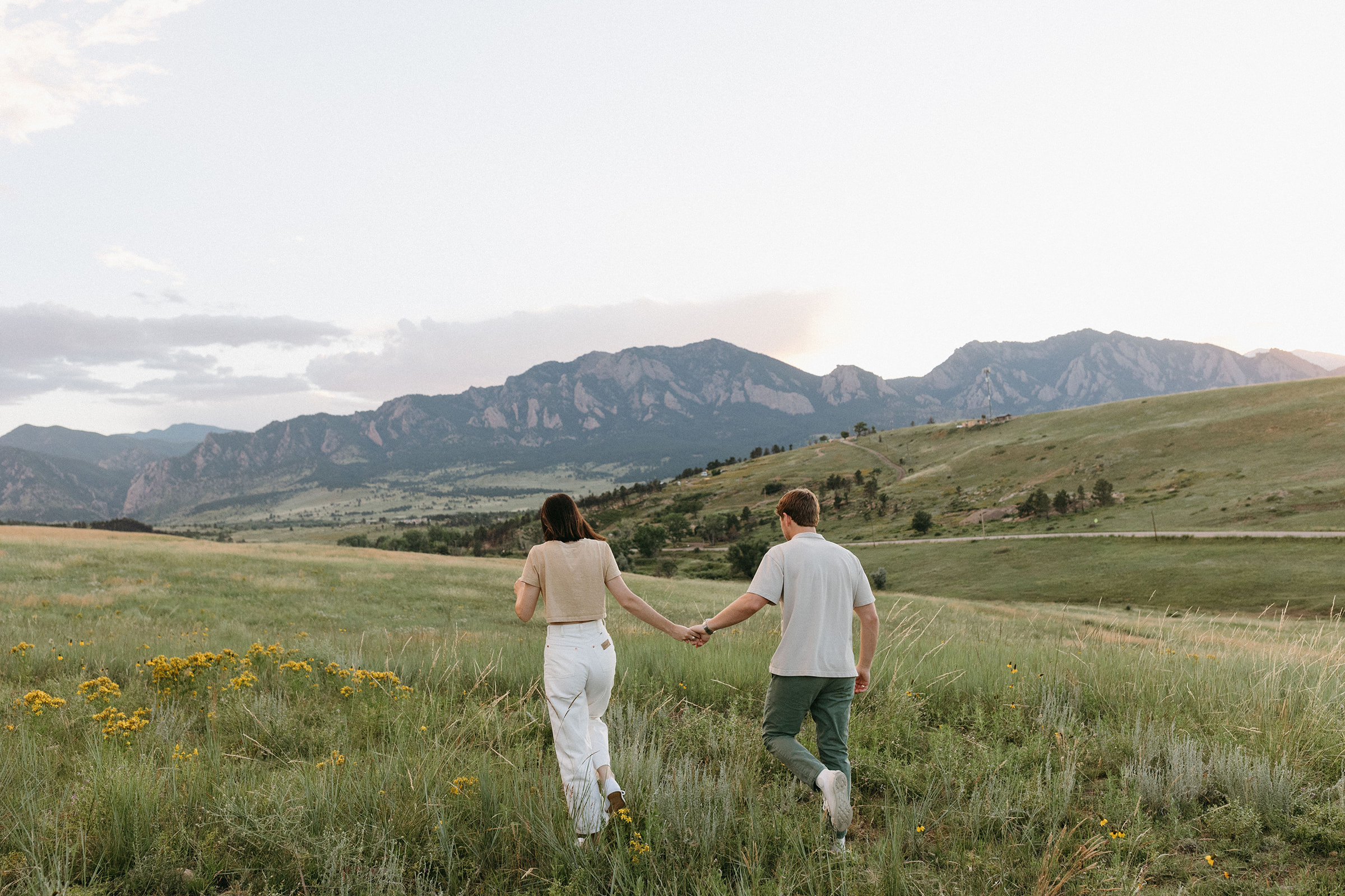 An editorial engagement session couple wearing natural-tone outfits walks through a wildflower meadow near Boulder
