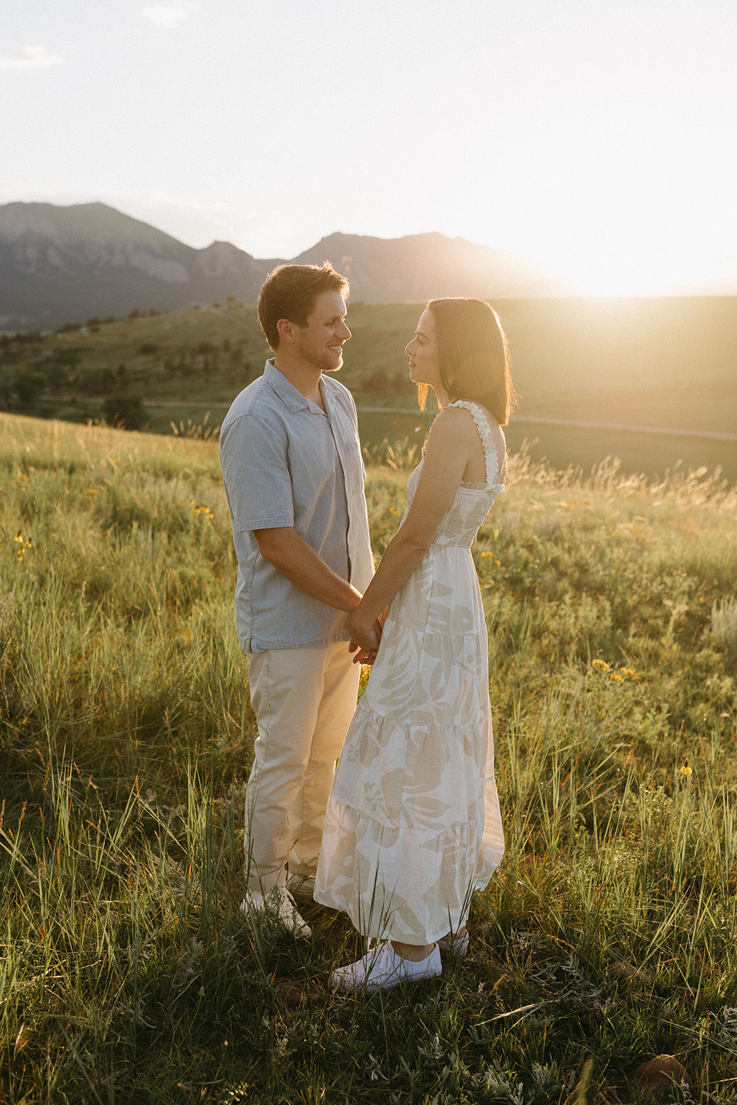A man wearing a denim, short-sleeved button-up shirt, and beige chino pants holds his fiancée's hands in a meadow. 