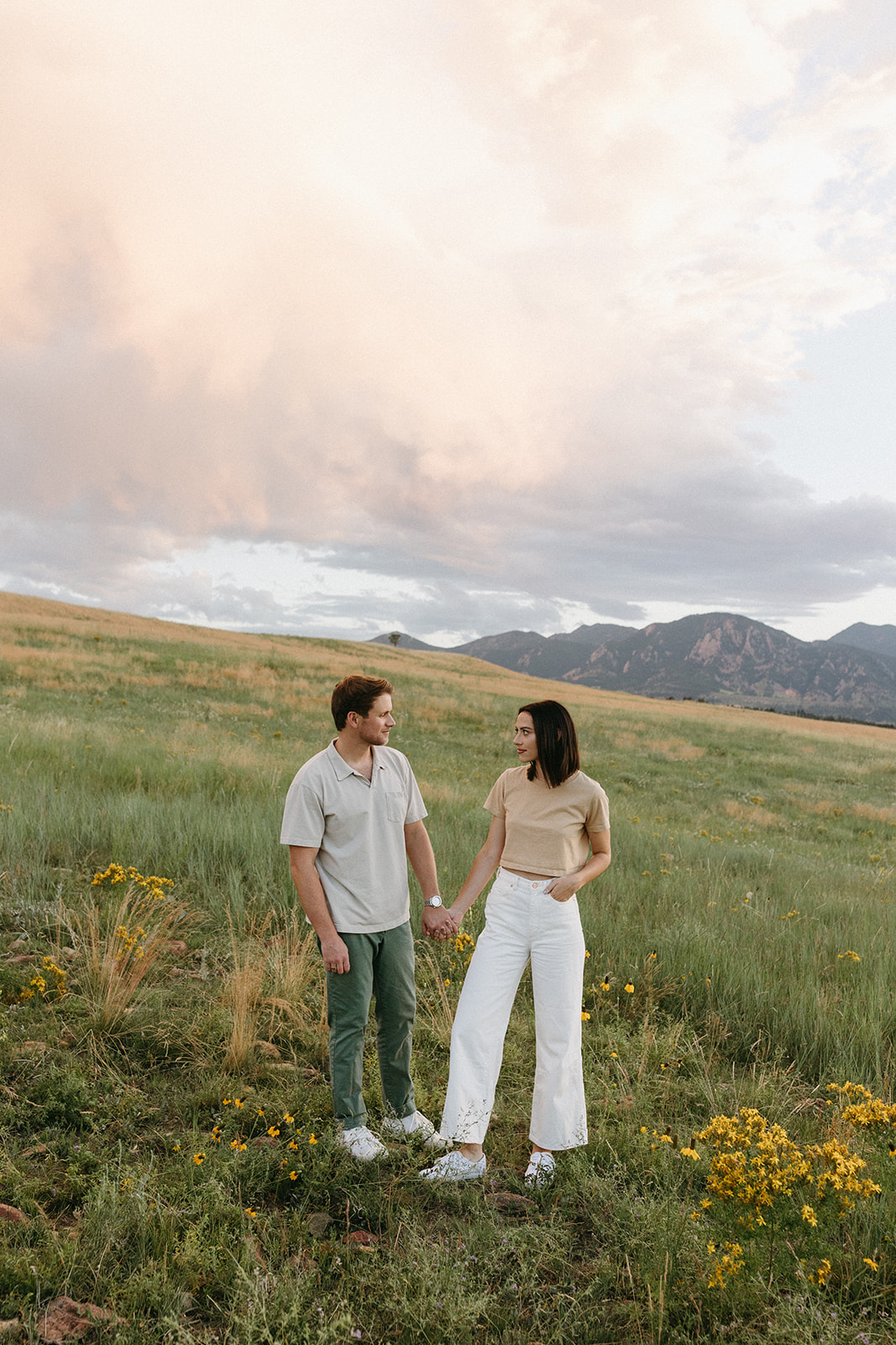 A Boulder engagement session couple holds hands while admiring one another in a wildflower meadow at Sunset in Colorado