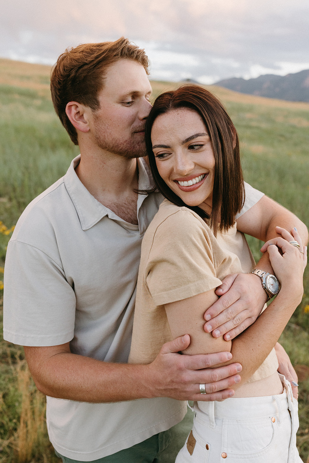 A stylish couple in neutral-tone outfits embraces in a meadow with a colorful Sunset and The Flatirons in the background