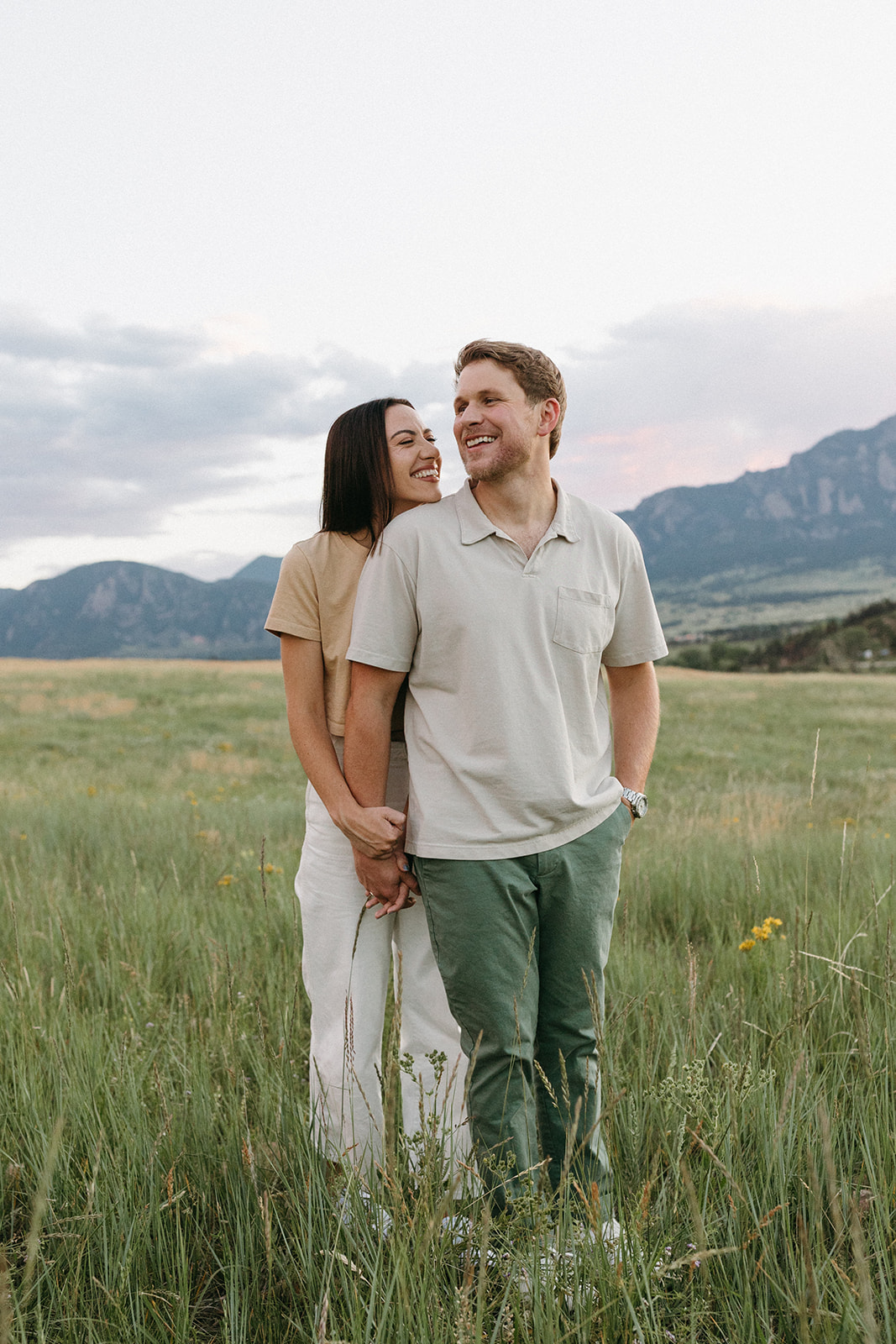A woman in a beige cropped tee and white jeans holds her fiancé's hand during a wildflower engagement session in Boulder