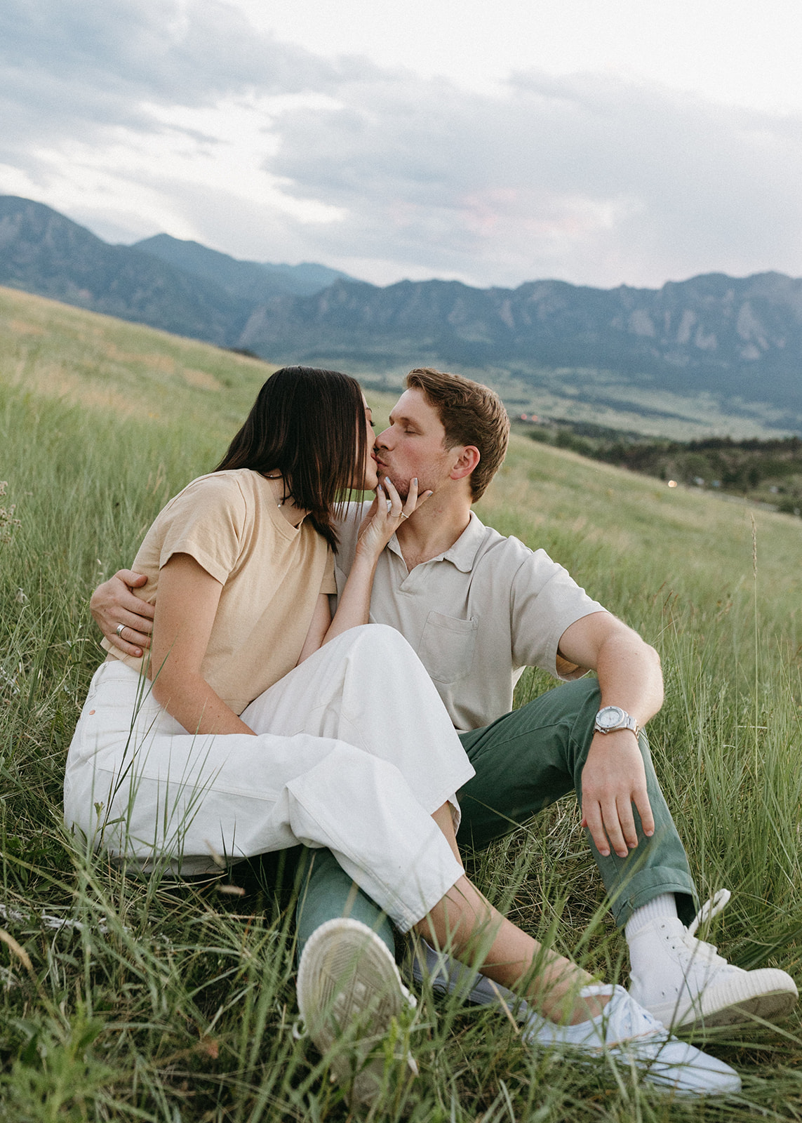An engaged couple in neutral attire kisses among colorful wildflowers, with Boulder's Flatirons in the background